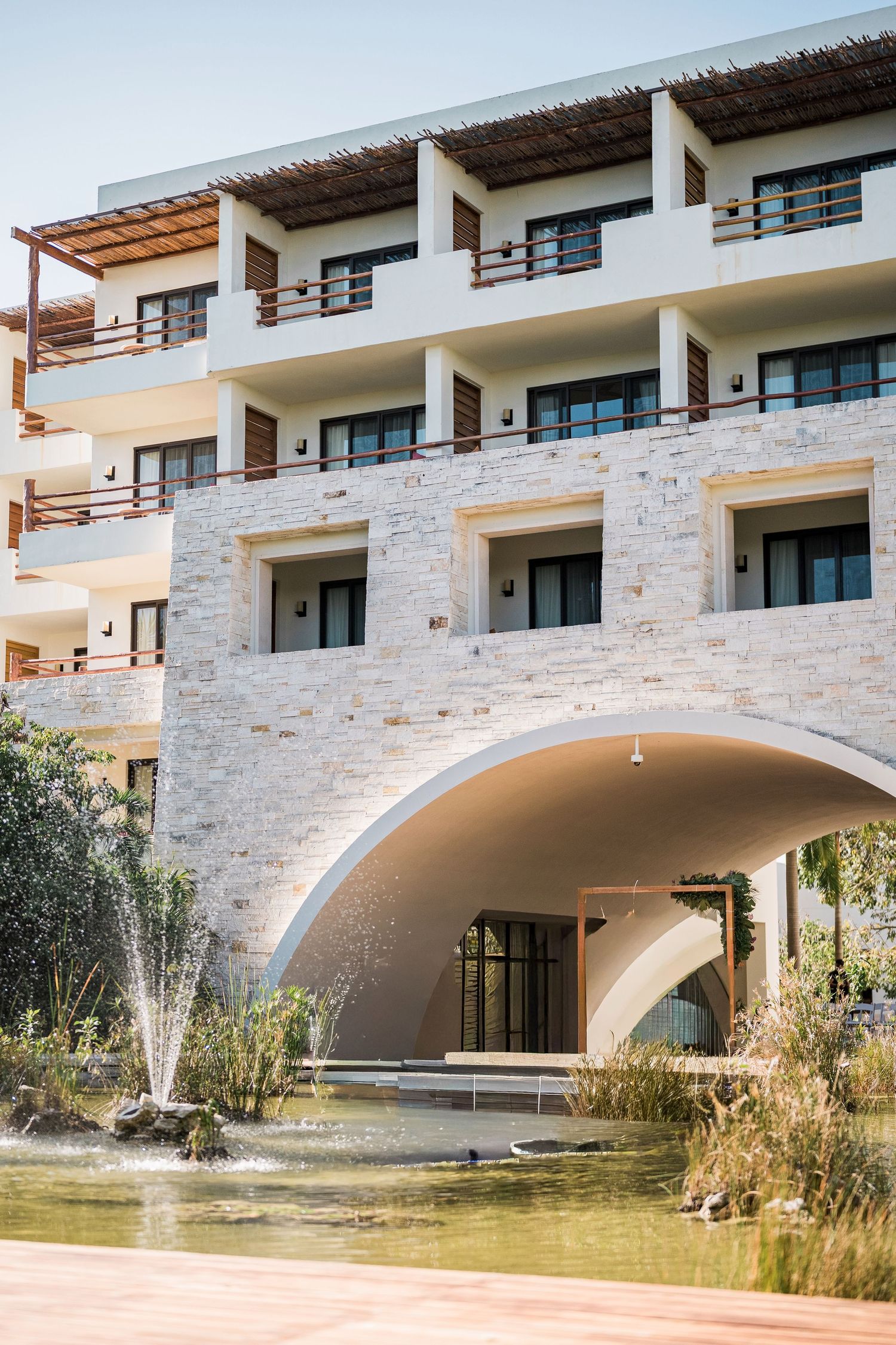 Secrets Akumal Wedding - Luxury resort with stone facade, arched entrance, fountain pond, and multi-story balconies under clear blue sky.