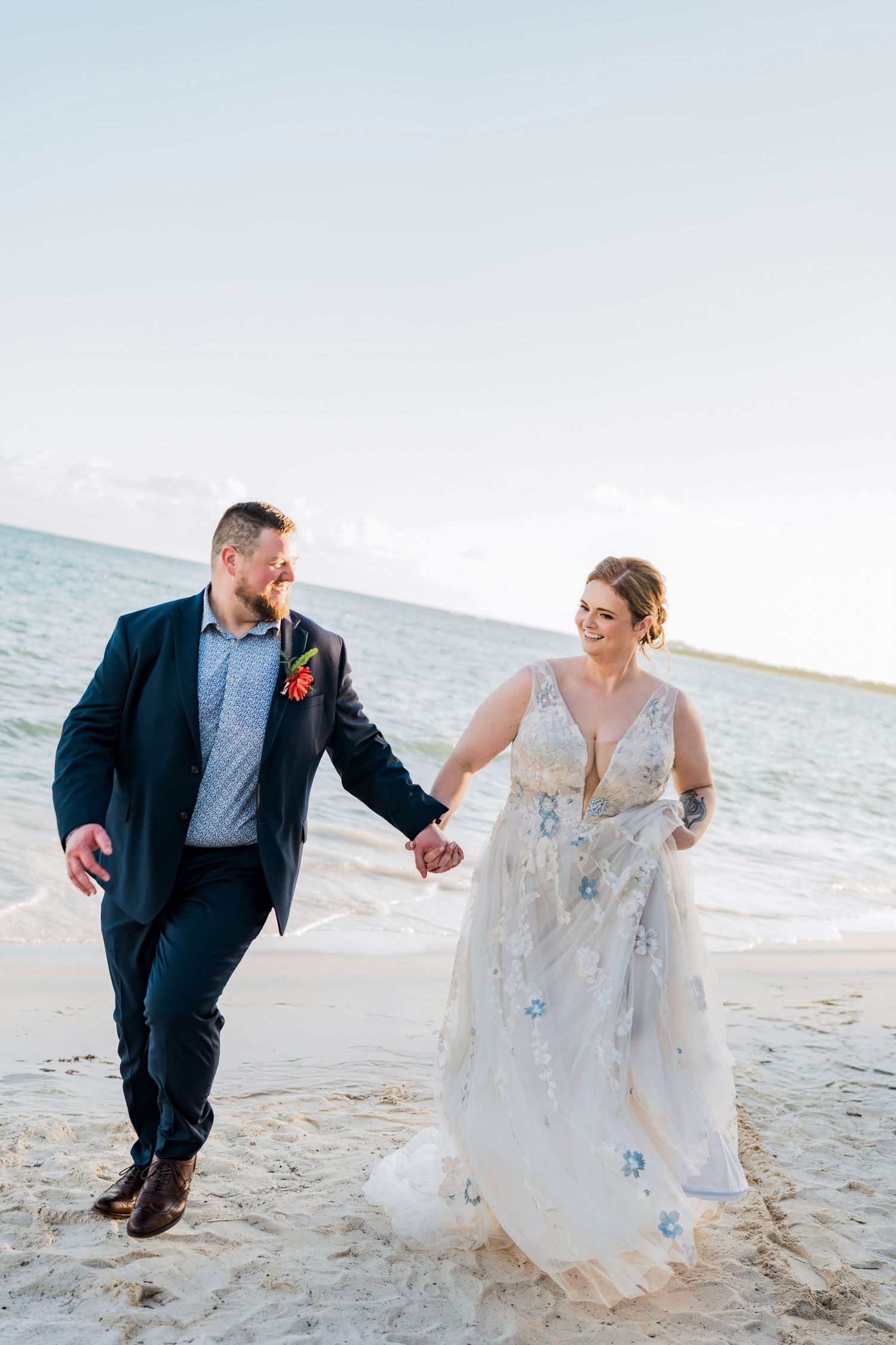 Secrets Akumal Wedding - A happy couple walks hand-in-hand on a sandy beach, the bride in a floral gown and groom in a navy suit.