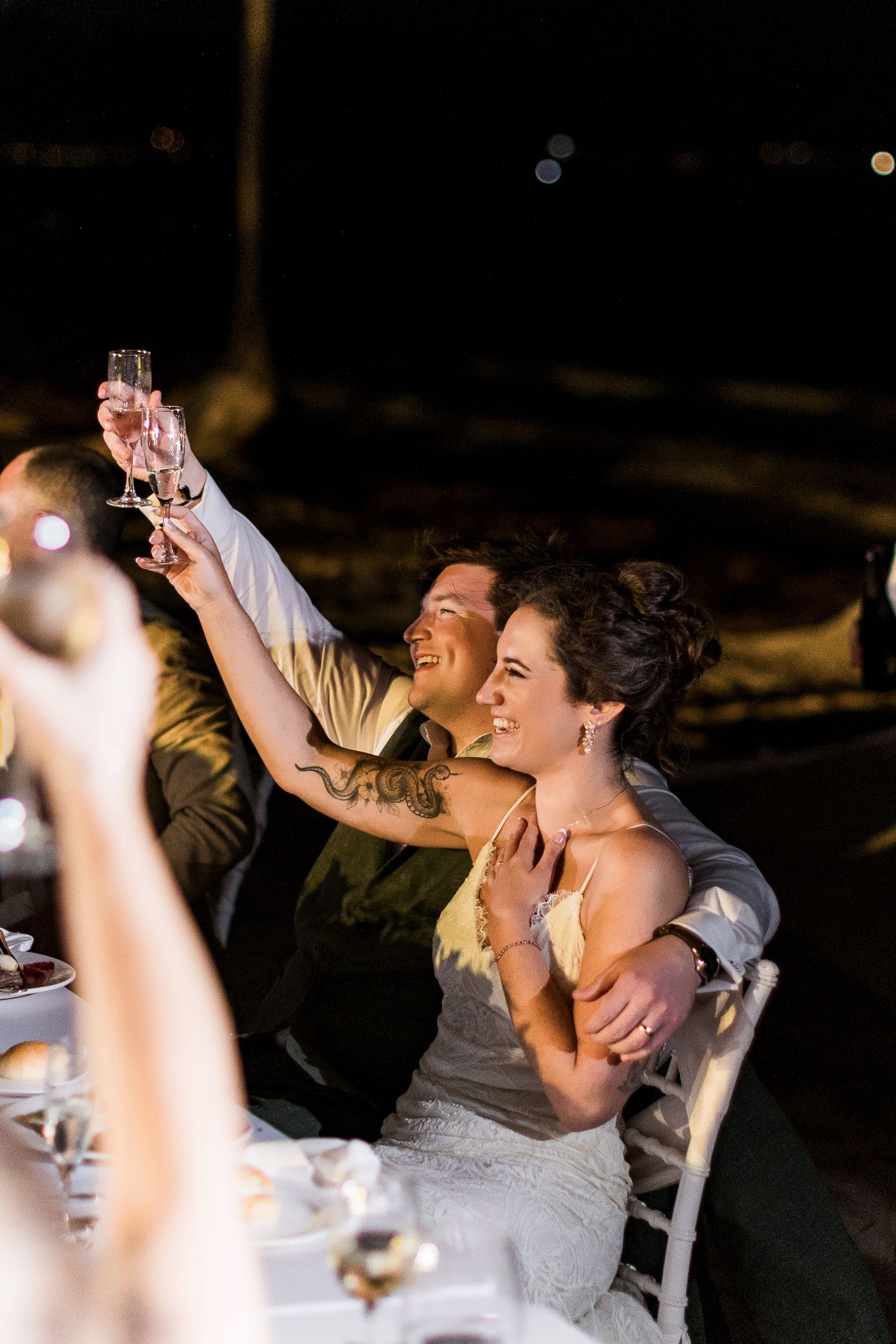 Bride and groom raising champagne glasses in a toast at their outdoor evening wedding reception dinner.