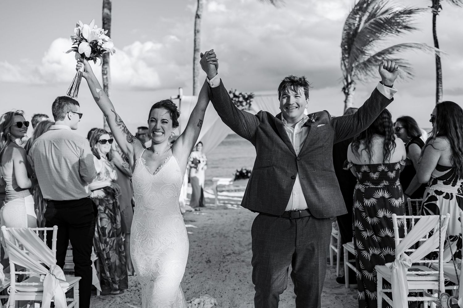 Joyful newlyweds celebrate at a beachside wedding ceremony, raising hands in triumph with guests cheering around them.