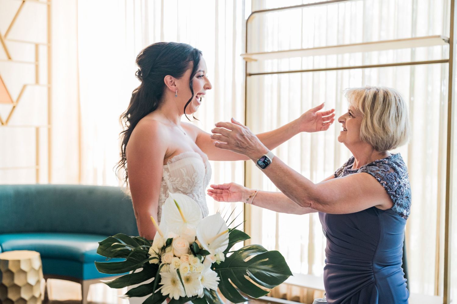 Mother helping bride prepare on wedding day, sharing a joyful moment in elegant bridal suite.