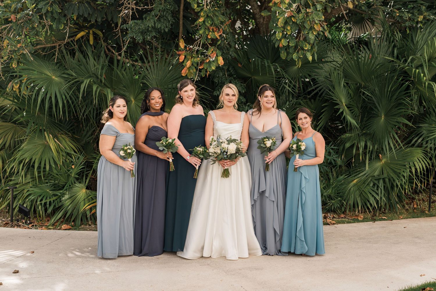 Bride in white gown poses with five bridesmaids in mismatched blue dresses, holding bouquets before lush tropical greenery.