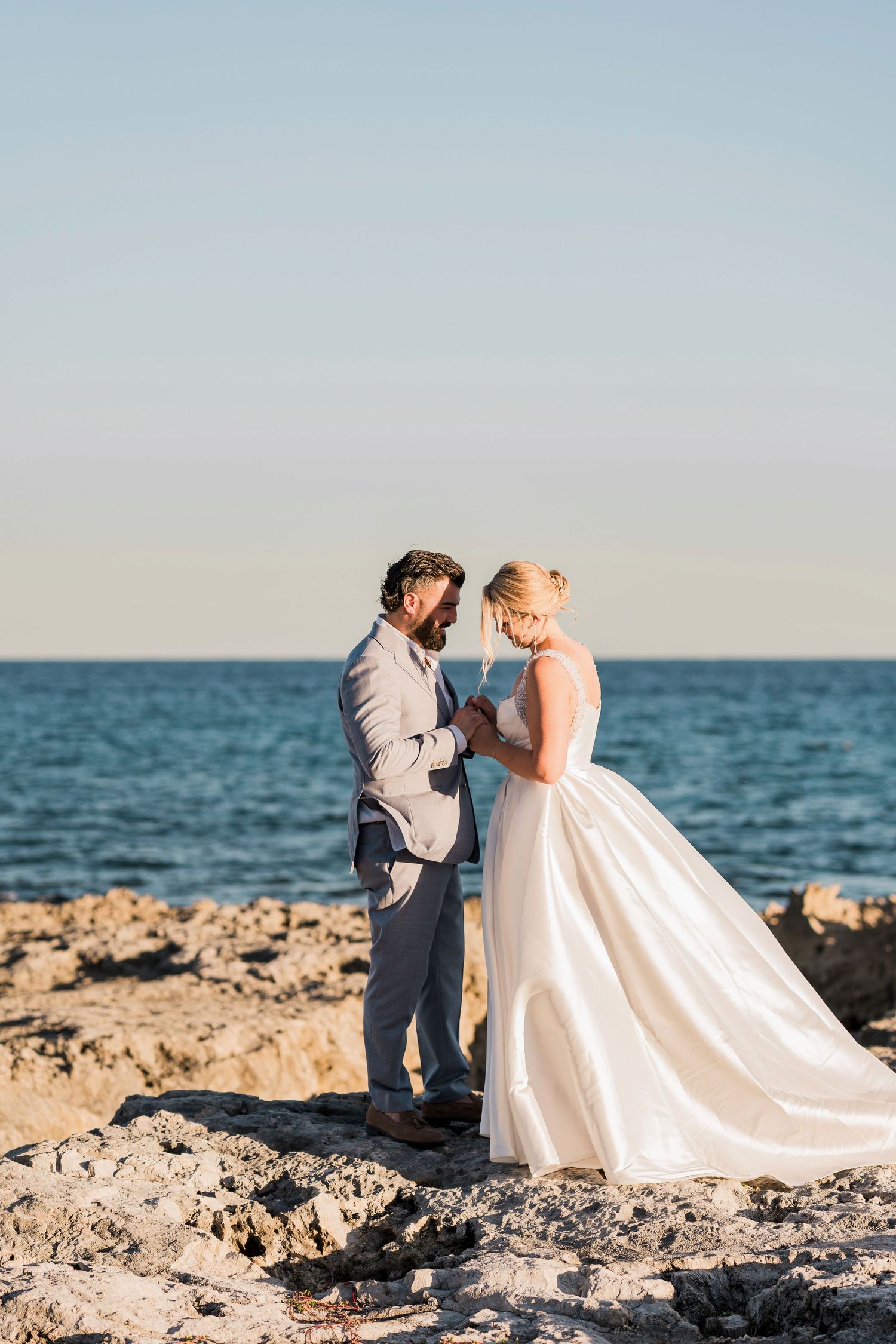 Bride and groom exchange vows on rocky shoreline at sunset, bride in white ballgown, groom in gray suit.