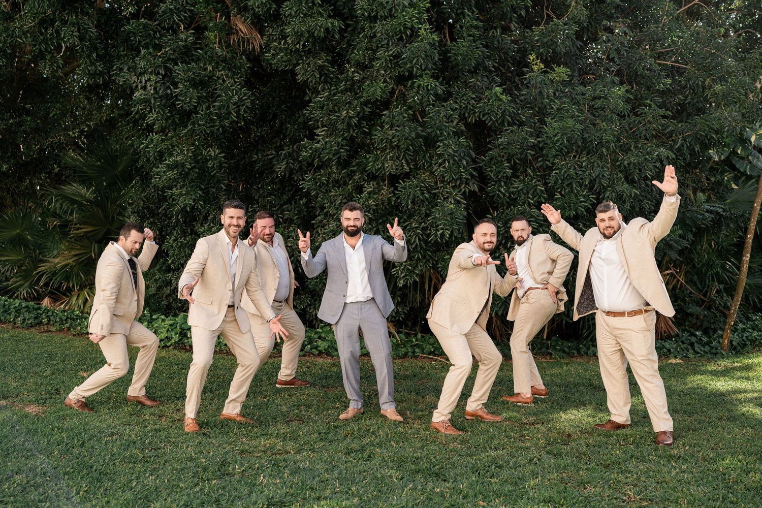 Groom in gray suit poses with groomsmen in tan suits on green lawn, all celebrating joyfully outdoors.