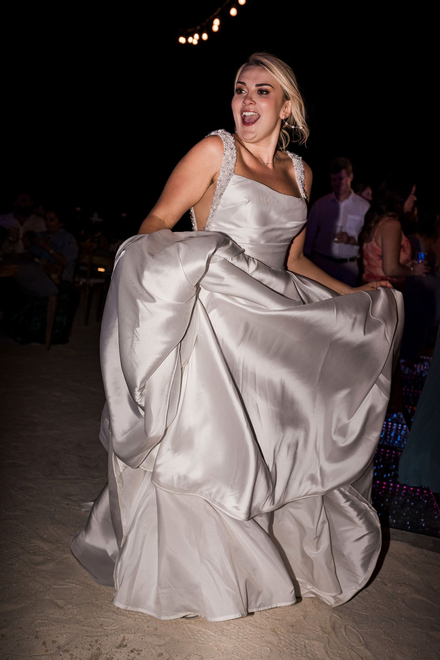 Woman in silver satin ball gown dancing joyfully on sandy beach at night outdoor wedding reception.
