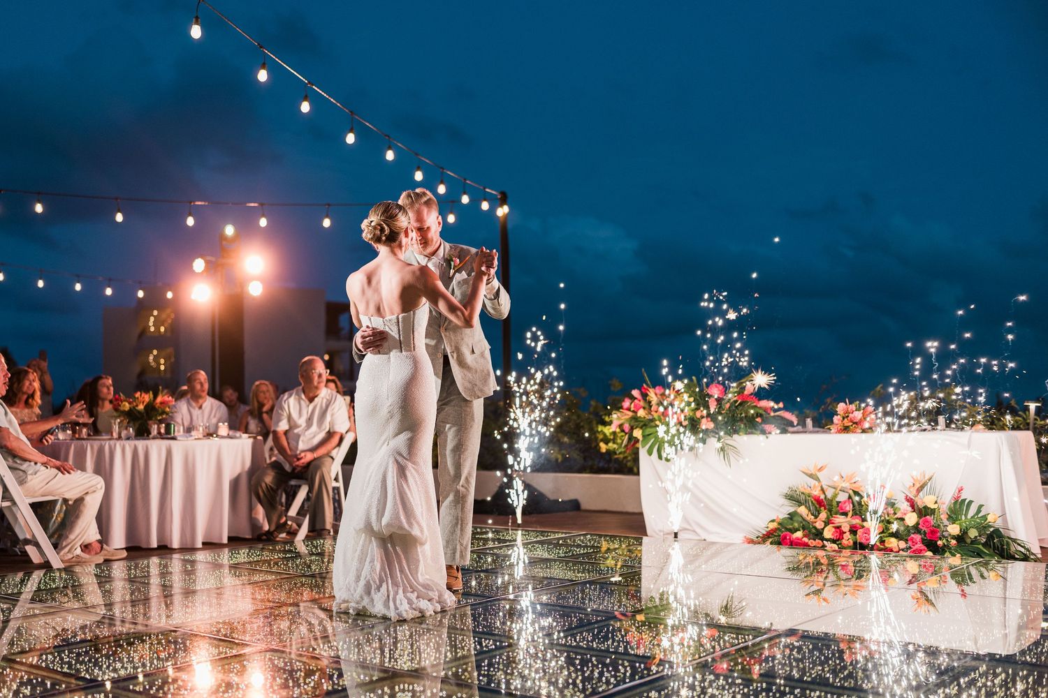 Couple shares first dance on reflective outdoor floor under string lights at elegant evening wedding reception.