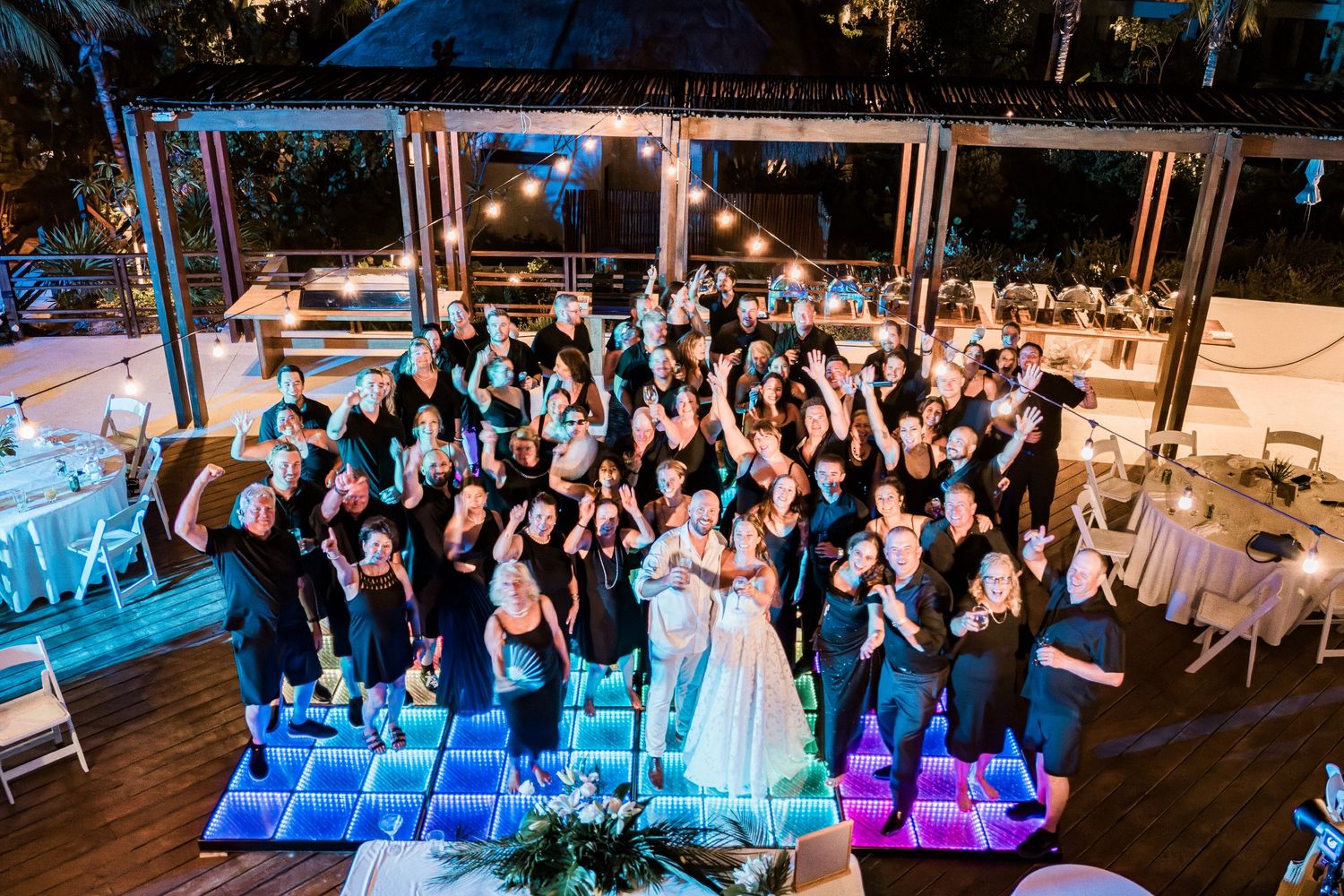 Aerial view of wedding party in black attire celebrating with bride on illuminated dance floor at outdoor venue.