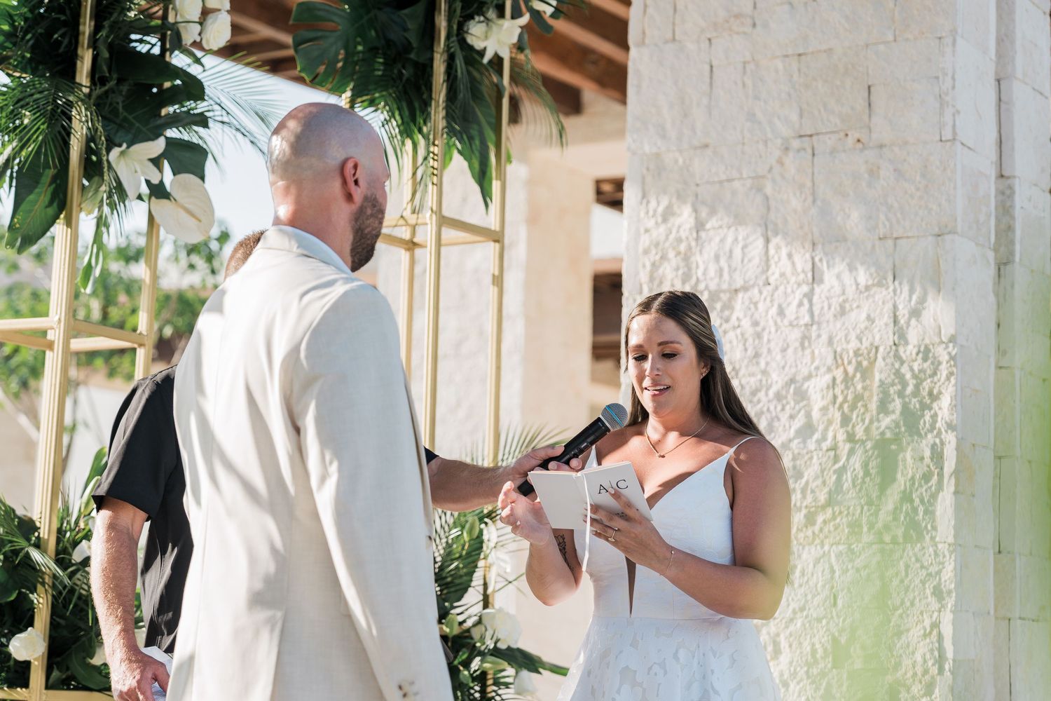 Bride reads vows to groom during outdoor wedding ceremony with tropical greenery backdrop.