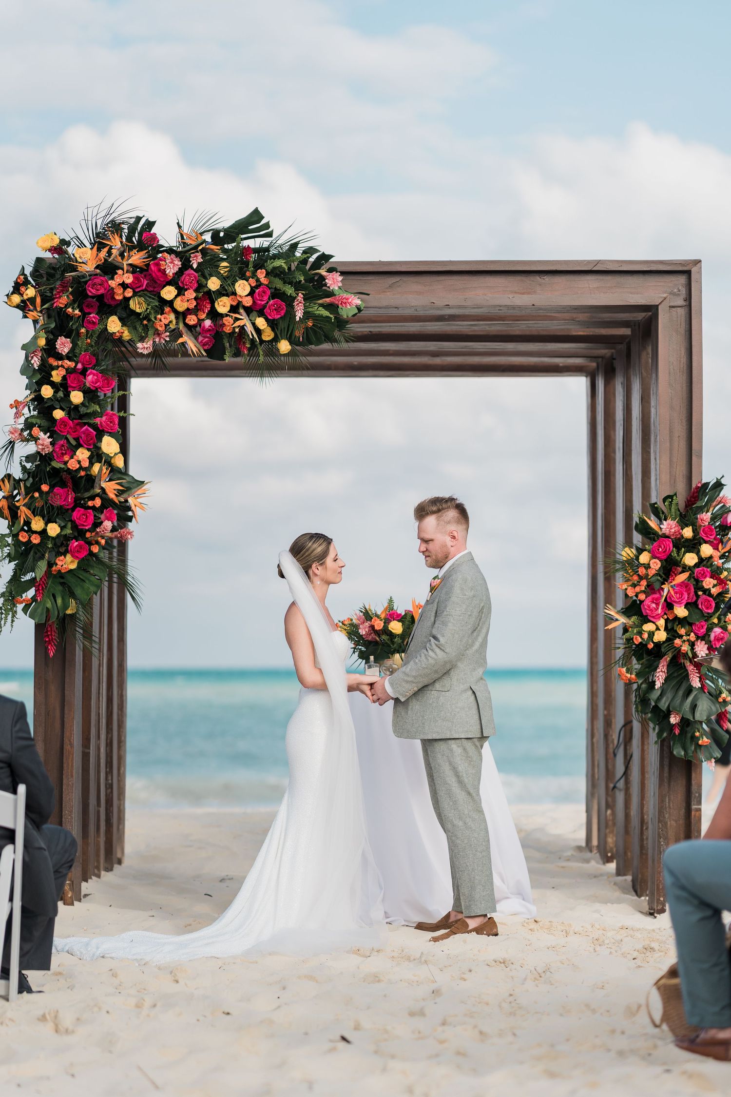 Bride and groom exchange vows under a floral arch on a tropical beach with turquoise ocean backdrop.