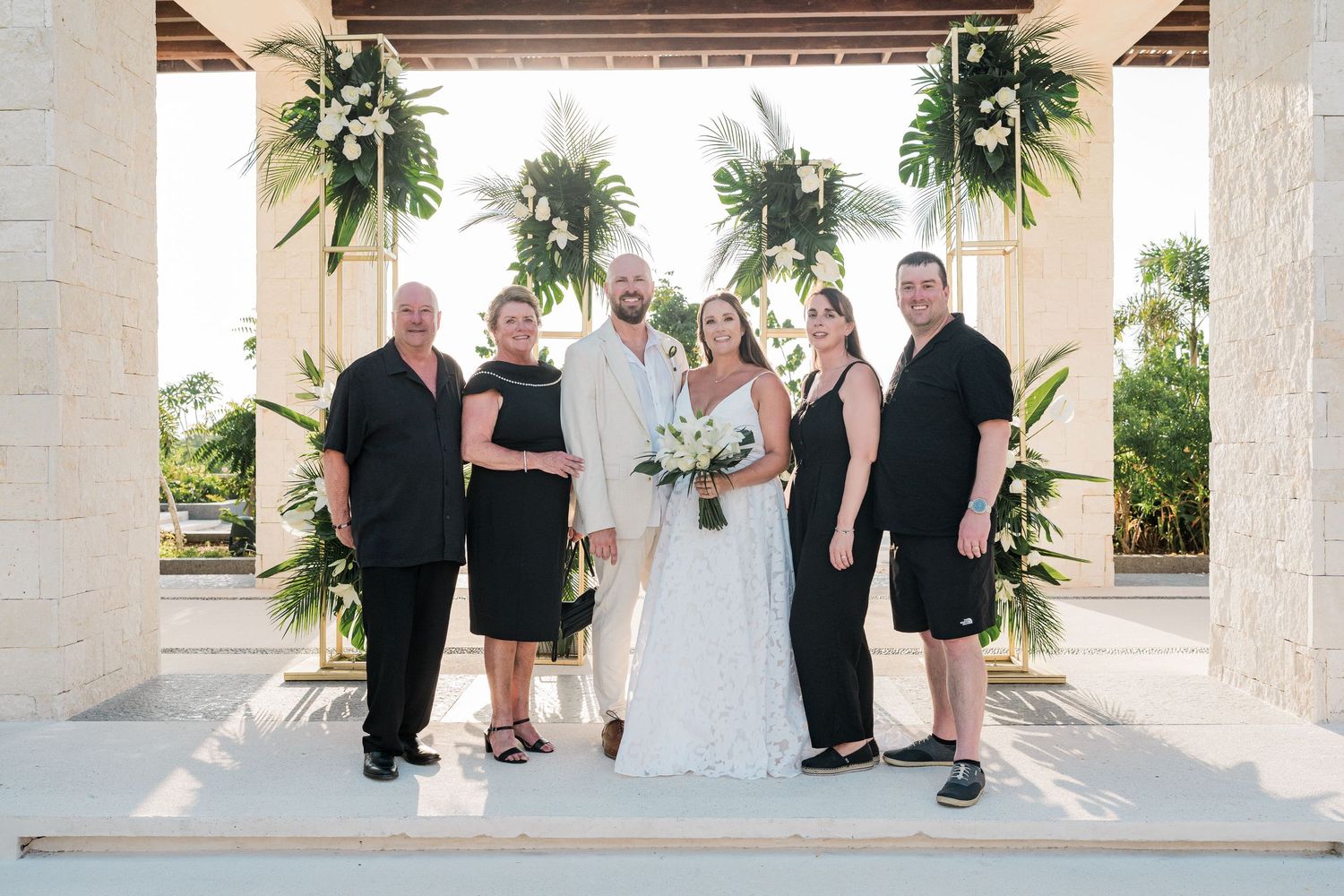 Wedding party poses under floral arch at tropical outdoor ceremony venue with lush greenery.