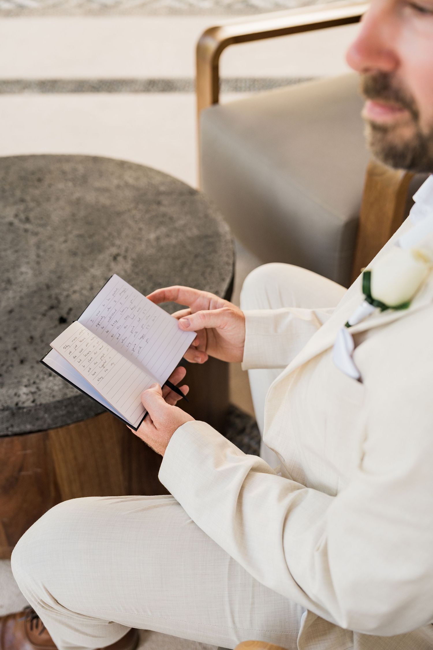 Groom in white suit reads handwritten wedding vows card while seated, wearing a white boutonniere.