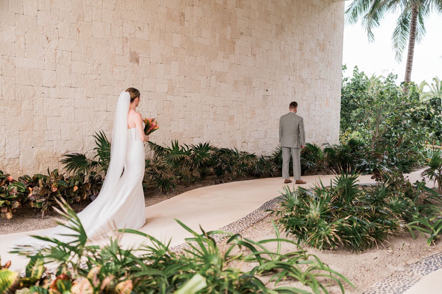Bride in white gown and veil approaches groom in gray suit for first look at tropical outdoor wedding venue.