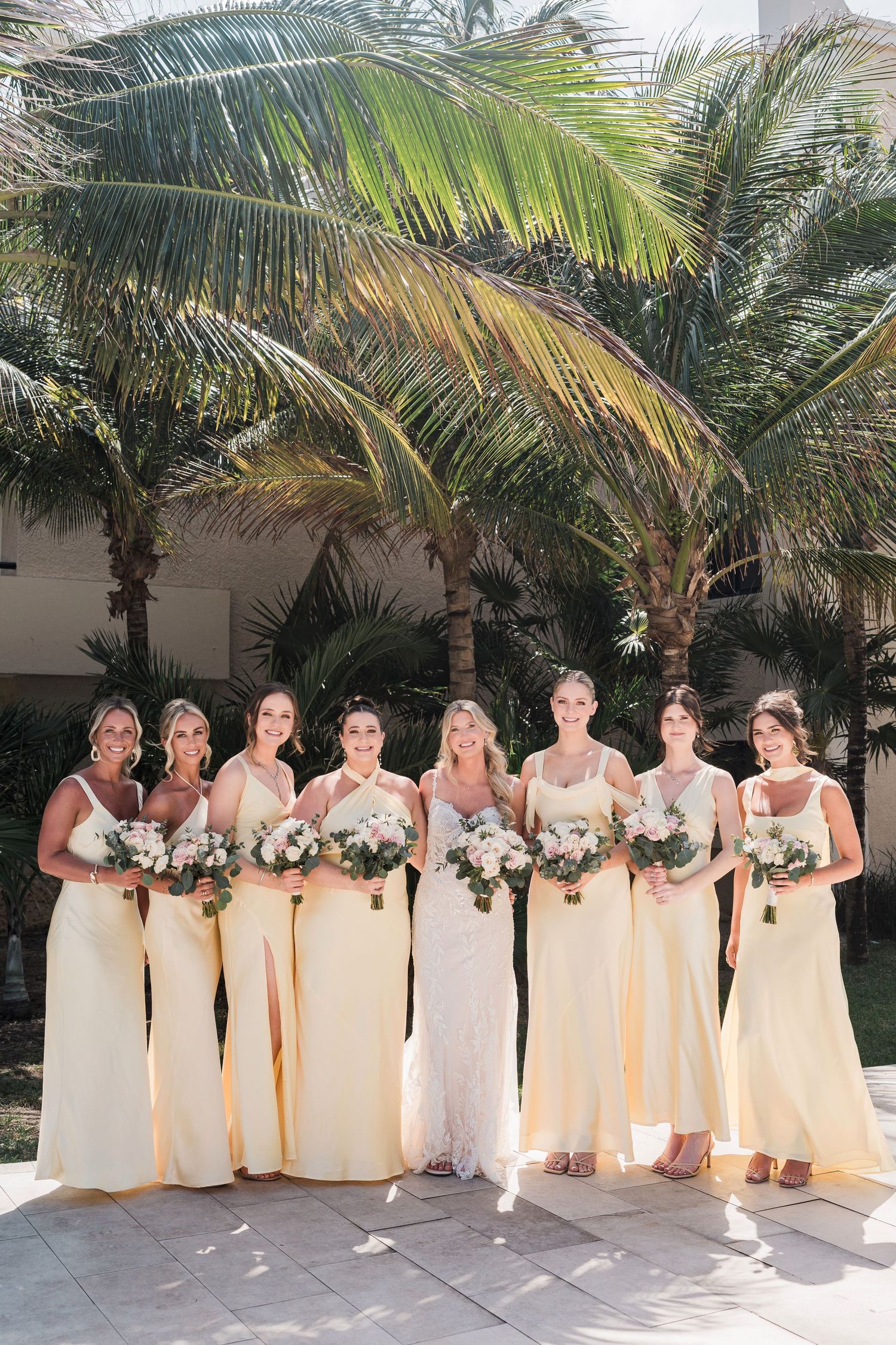 Bride and six bridesmaids in yellow gowns holding floral bouquets pose before tropical palm trees at an outdoor wedding.