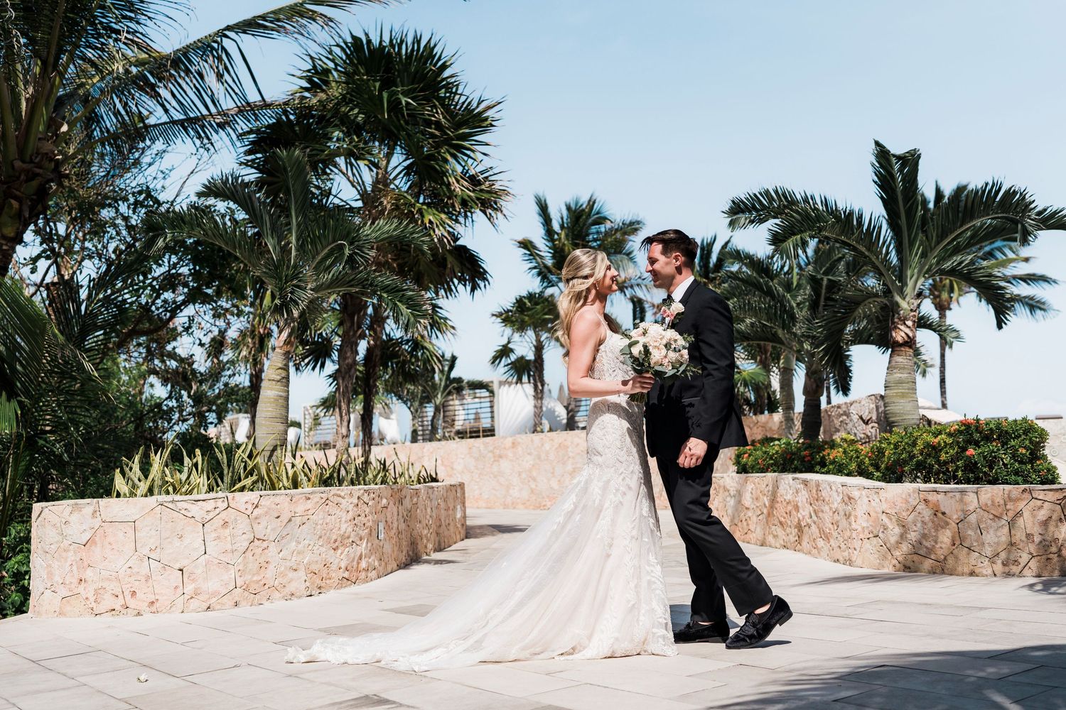 Bride and groom share a romantic moment on a tropical terrace surrounded by palm trees in a destination wedding setting.
