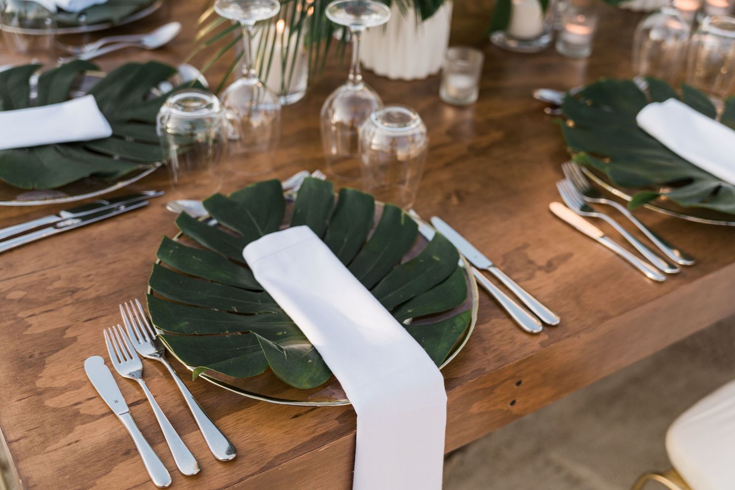 Elegant tropical wedding table setting with monstera leaf chargers, white napkins, and silver cutlery on a wooden table.