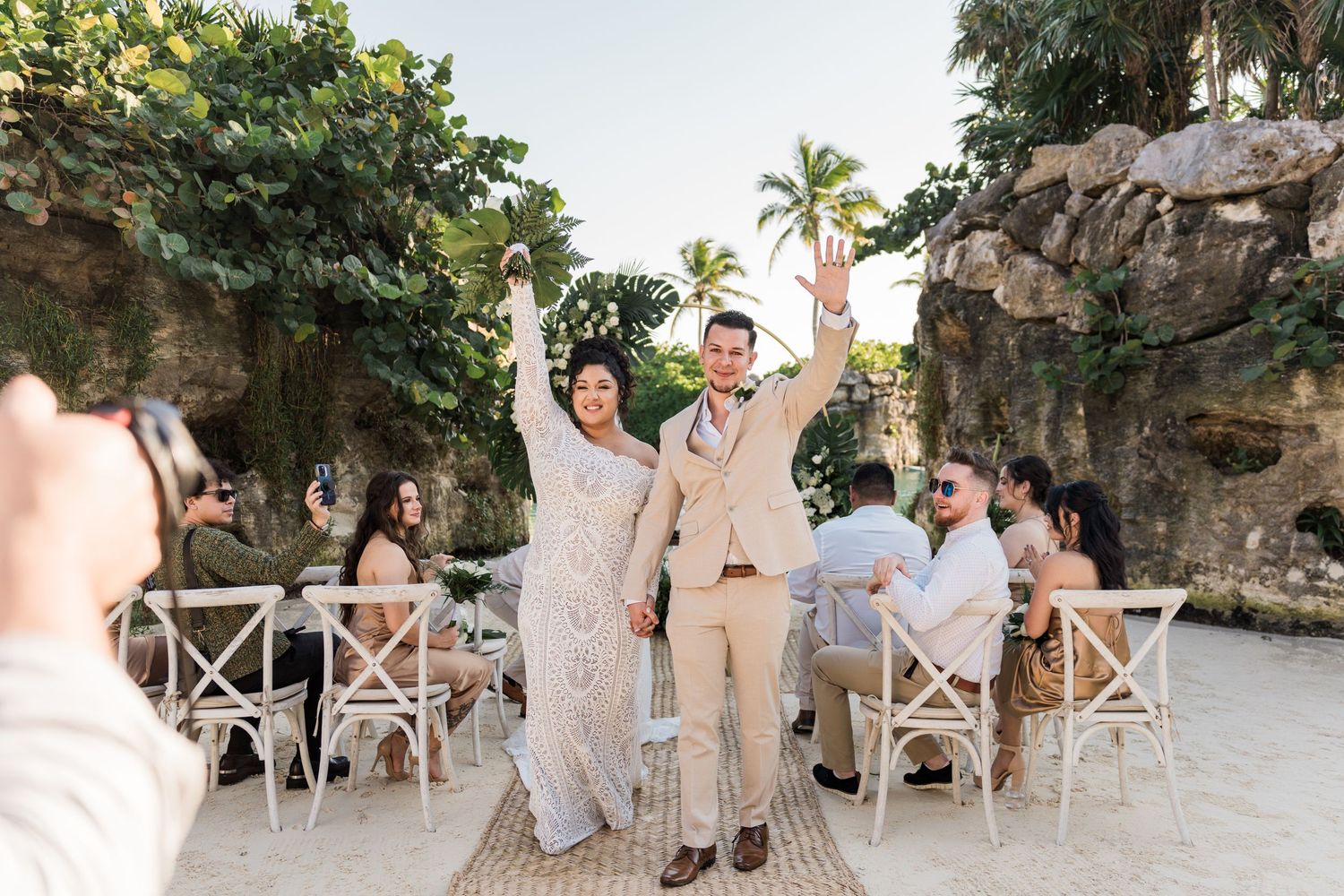 Xcaret Arte Wedding - Bride and groom raise hands joyfully at outdoor beach wedding ceremony with guests seated among tropical rocks.