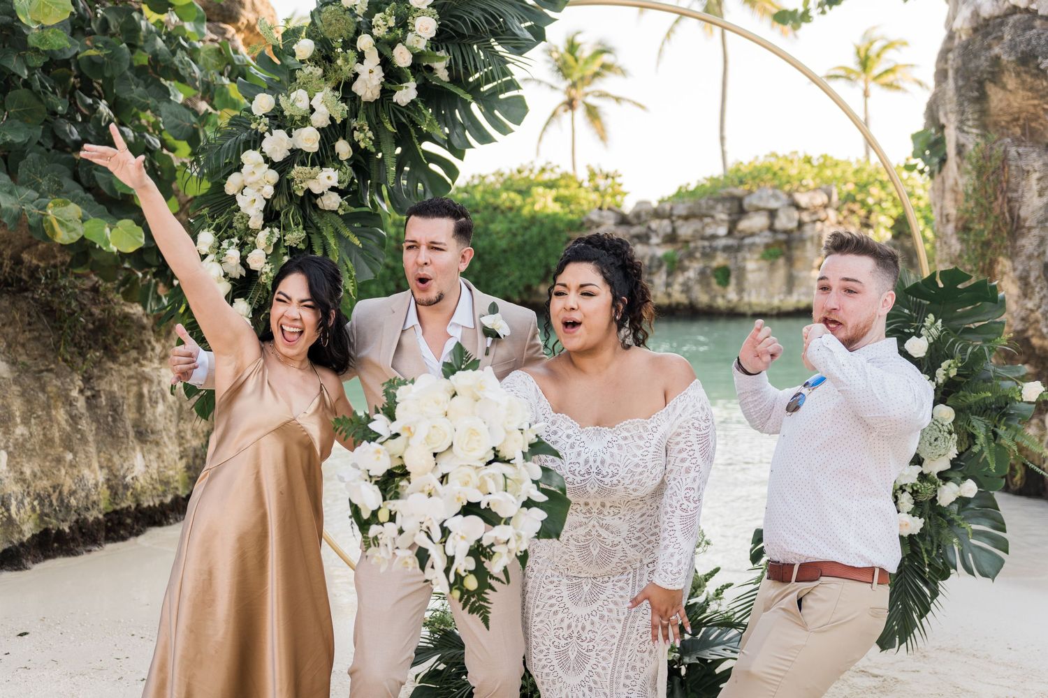Xcaret Arte Wedding - Joyful wedding party celebrating on a tropical beach with white floral arch, bride in lace gown holding bouquet.