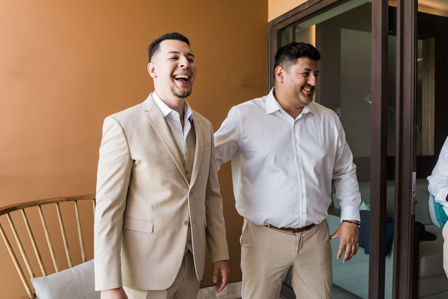Xcaret Arte Wedding - Two men in smart casual attire laugh together, one wearing a beige suit, in a bright indoor setting.