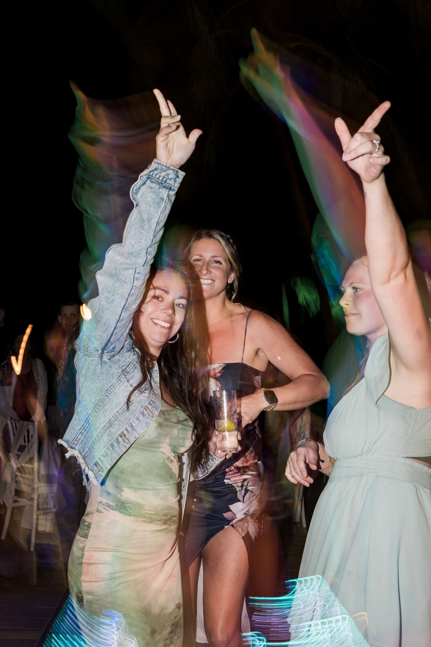 Three women dancing and celebrating at a nighttime outdoor party, making rock on hand gestures under colorful lights.