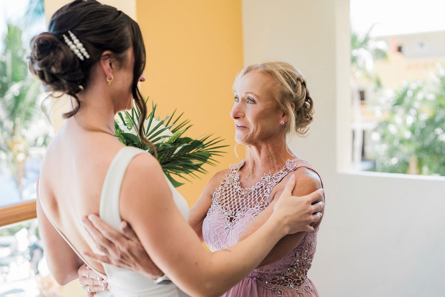 Bride in white gown embraces older woman in lavender dress, sharing tender moment at tropical wedding venue.