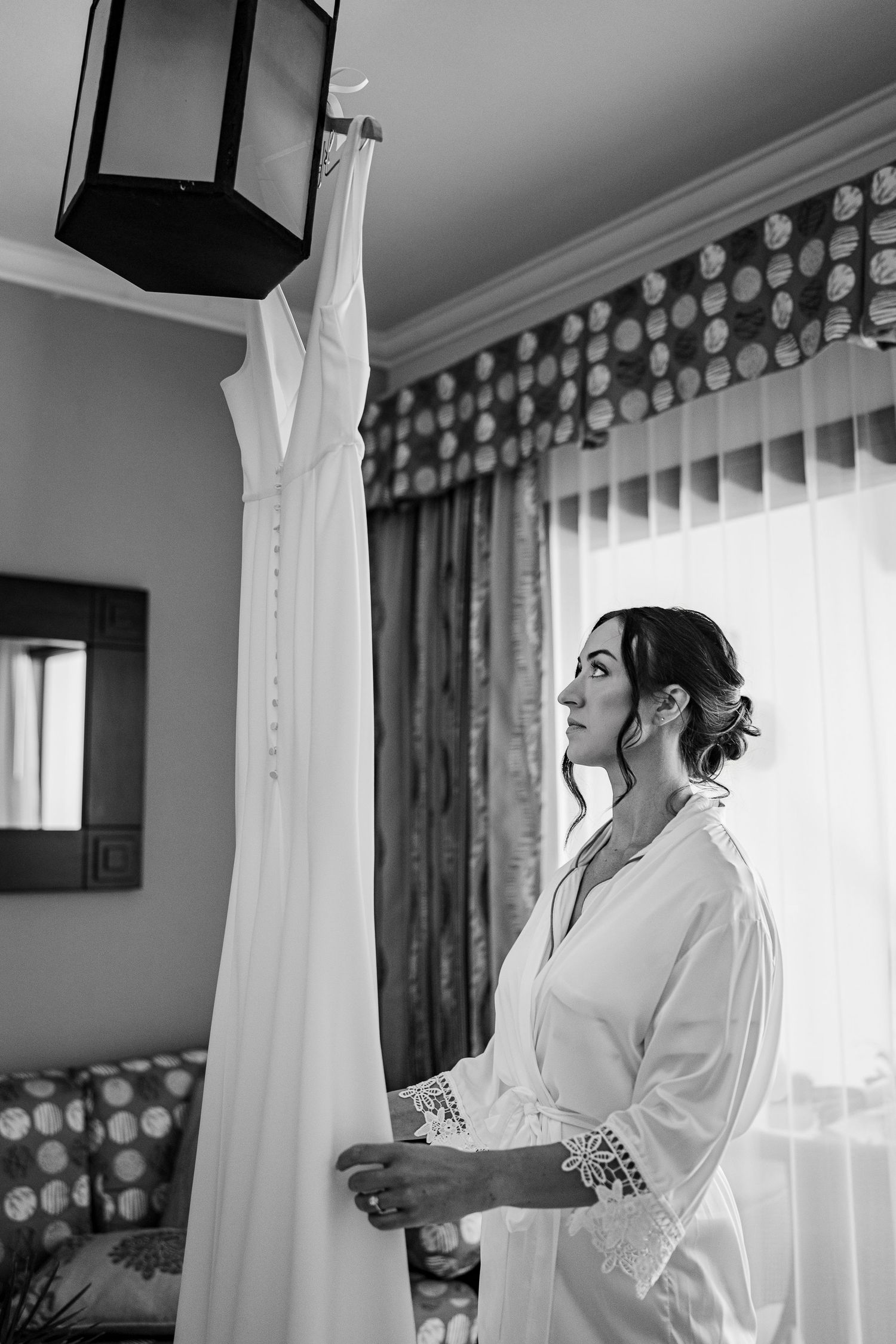 Bride in white robe admiring her wedding dress hanging in a sunlit room, black and white photo.