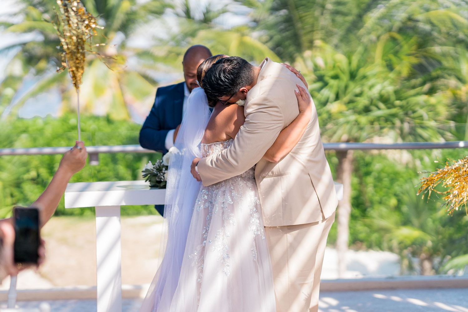 Bride and groom share an emotional hug during their outdoor beach wedding ceremony with palm trees in background.