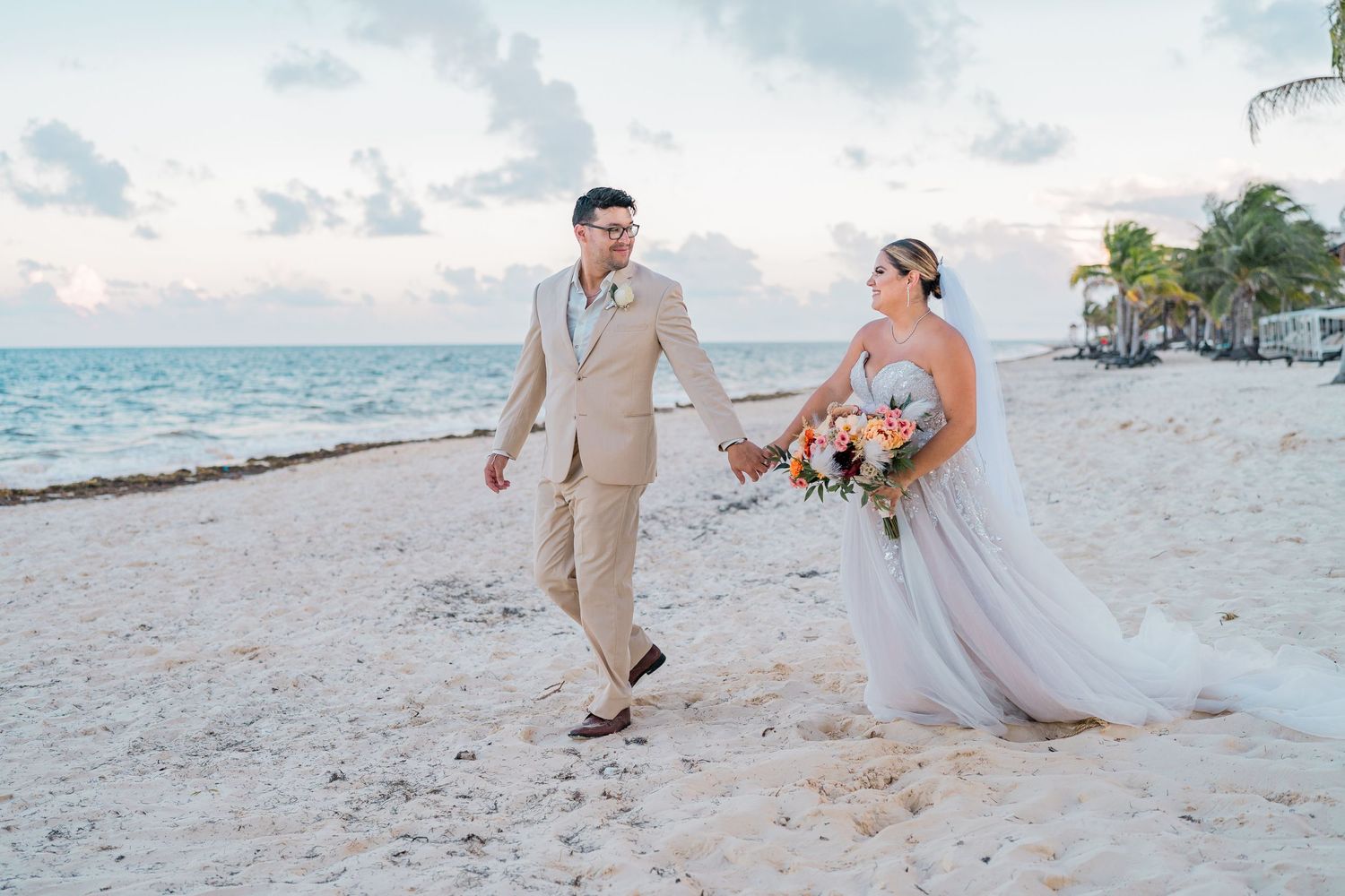 Bride and groom walking hand in hand along a white sand tropical beach at dusk.