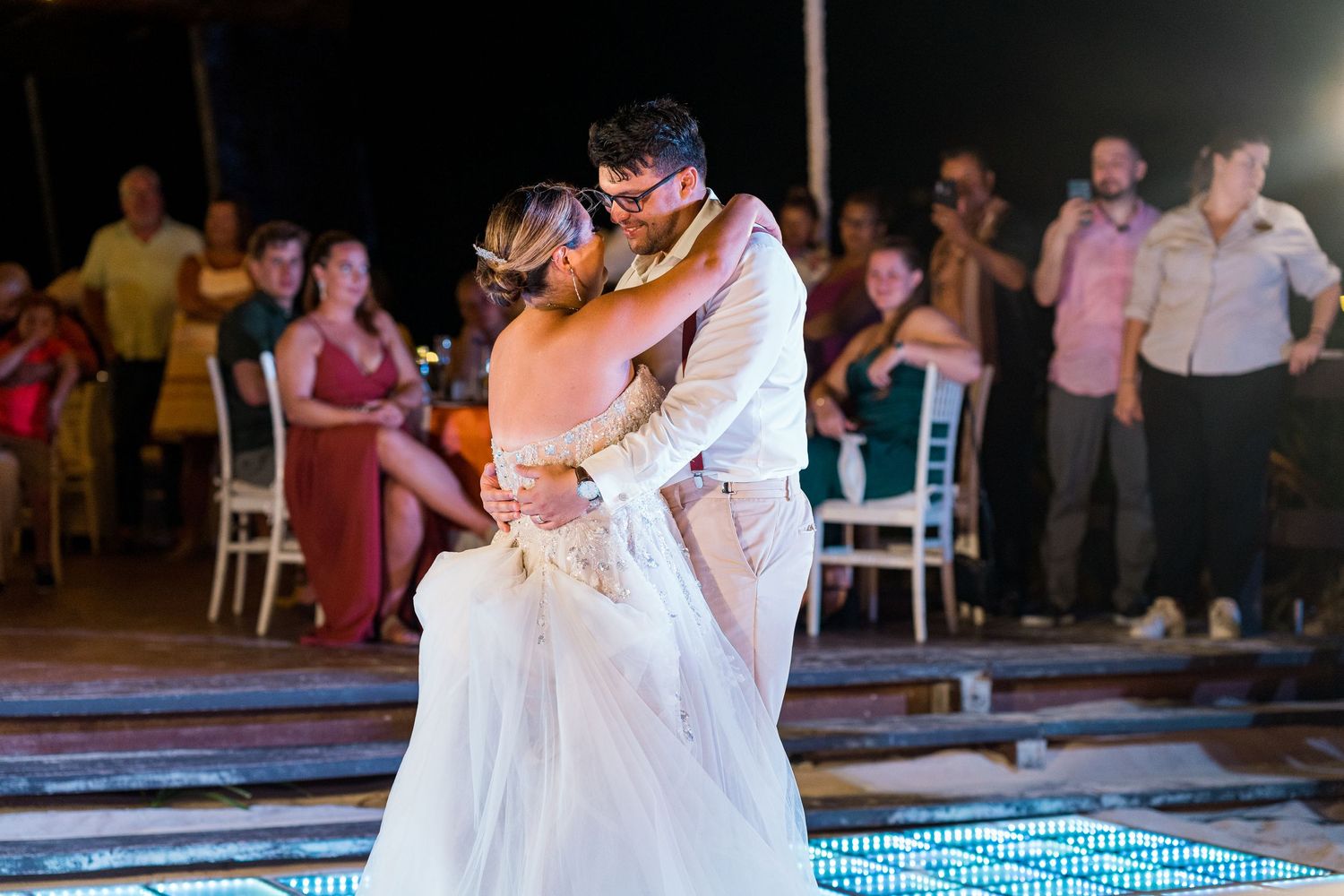 Bride and groom share first dance surrounded by wedding guests on illuminated dance floor.