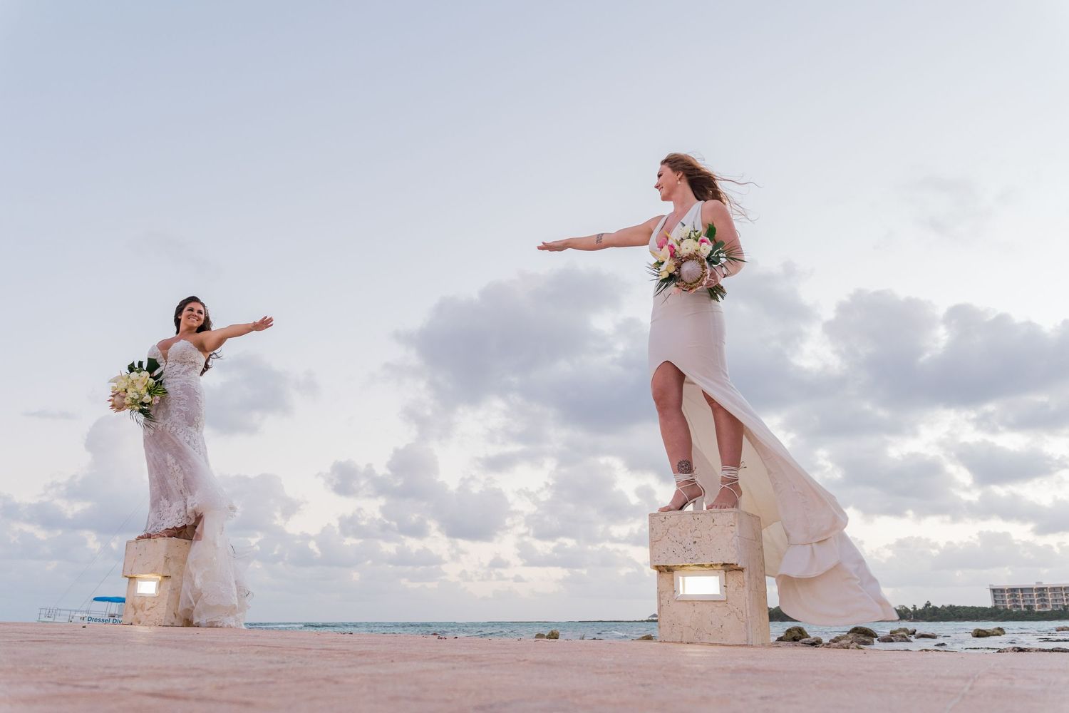Two brides playfully posing with arms outstretched on stone pillars by the ocean.