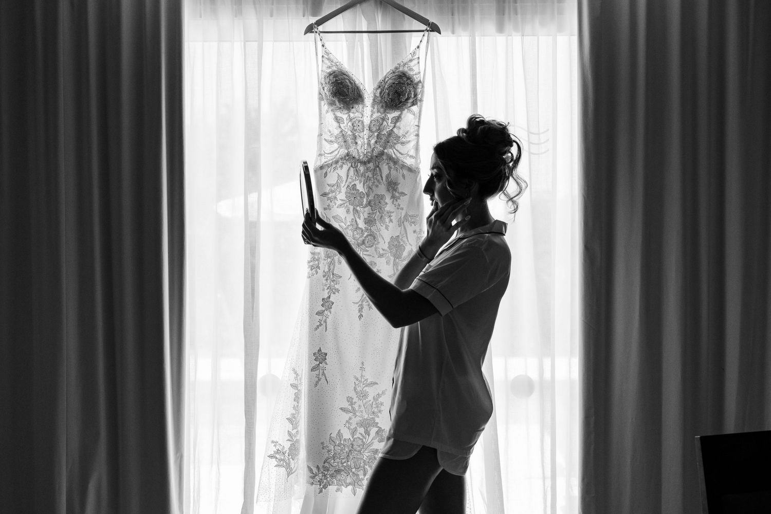 Black and white silhouette of bride holding tablet next to her hanging beaded wedding gown.