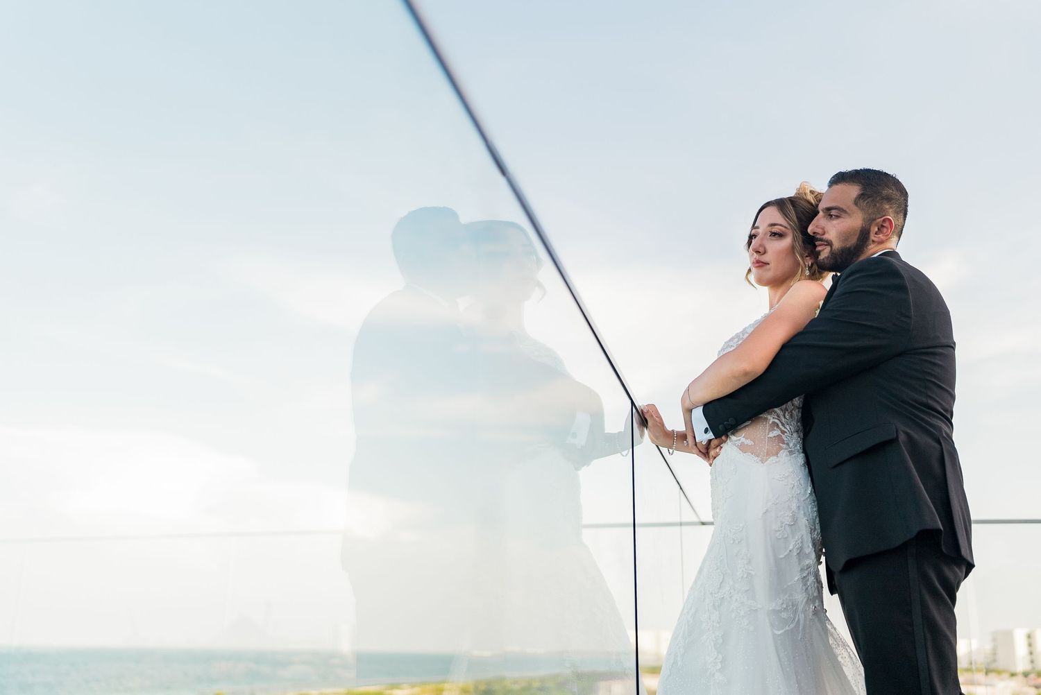Bride and groom share a romantic embrace on a glass balcony overlooking the ocean on their wedding day.