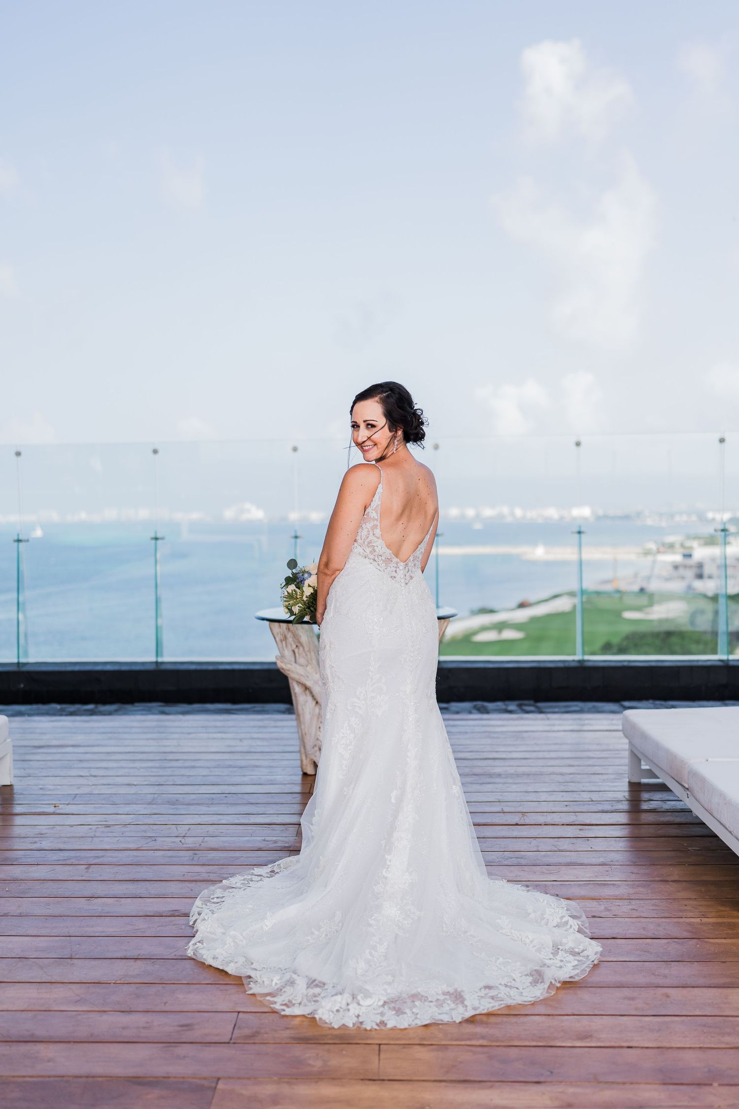 Bride in lace backless gown poses on rooftop terrace with ocean view backdrop.