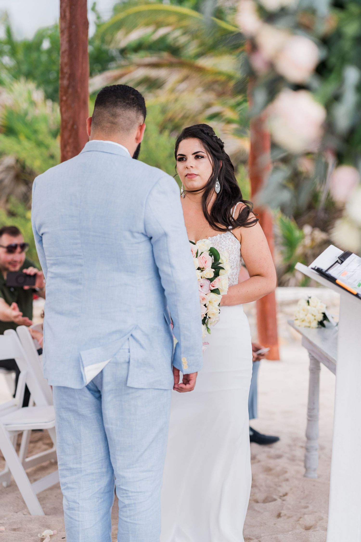 Bride in white gown holds bouquet while groom in light blue suit holds hands during beachside wedding ceremony.