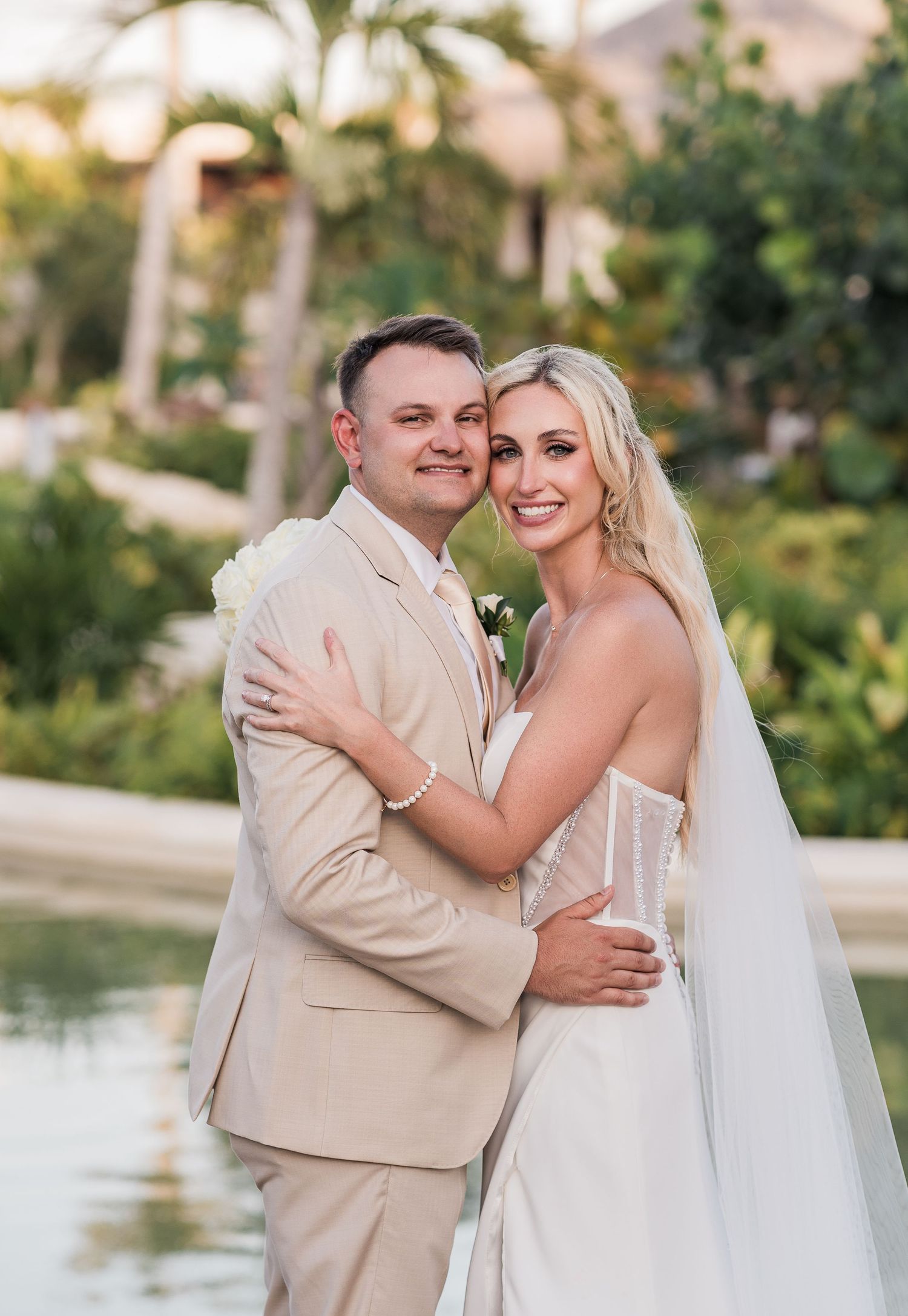 Smiling bride and groom pose together near a tropical resort pool at golden hour.