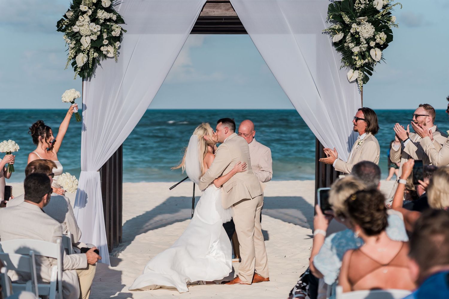 Newlyweds share first kiss under white draped floral arch at beach wedding as guests applaud.