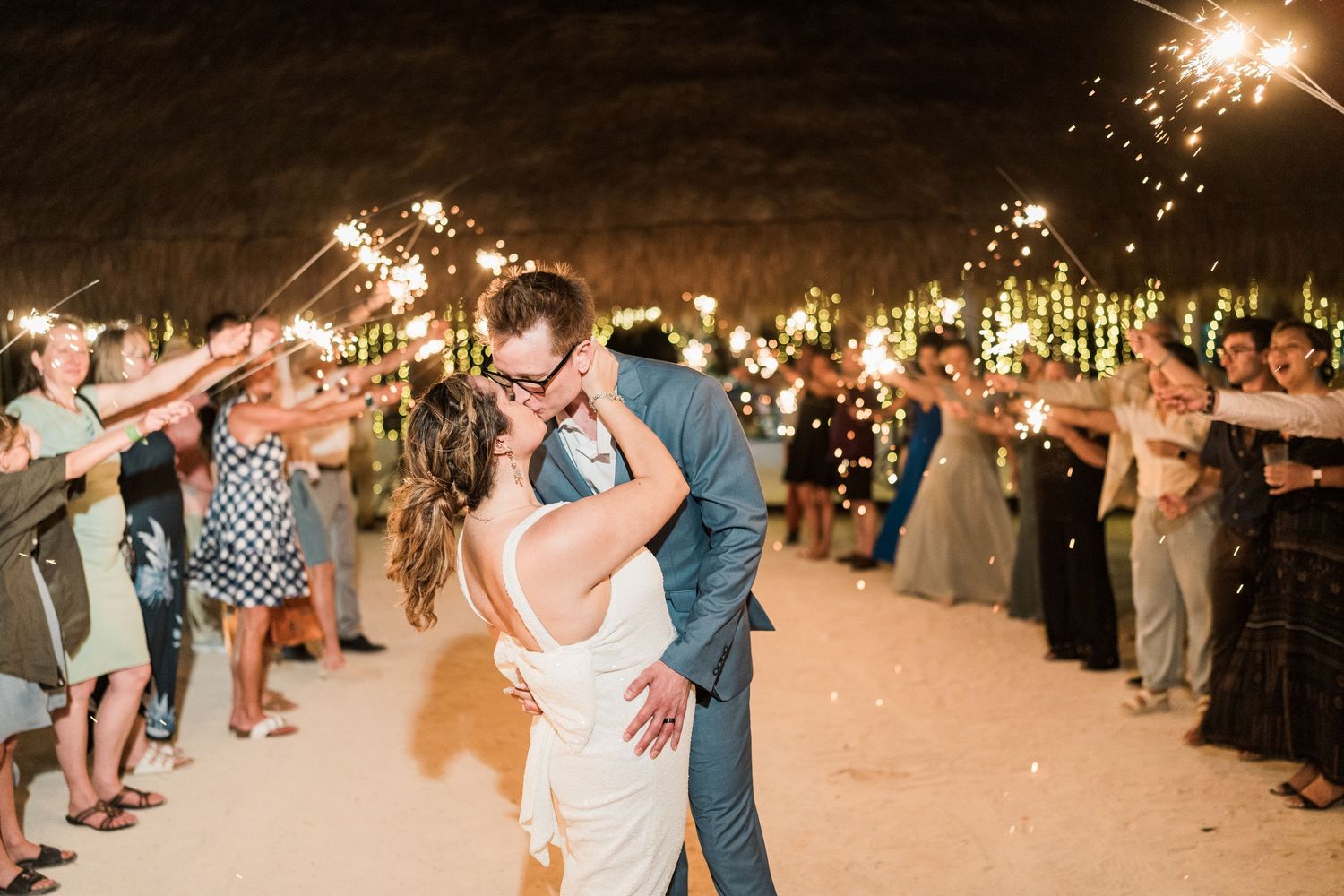 Bride and groom sharing romantic kiss during sparkler send-off at beachside wedding reception.