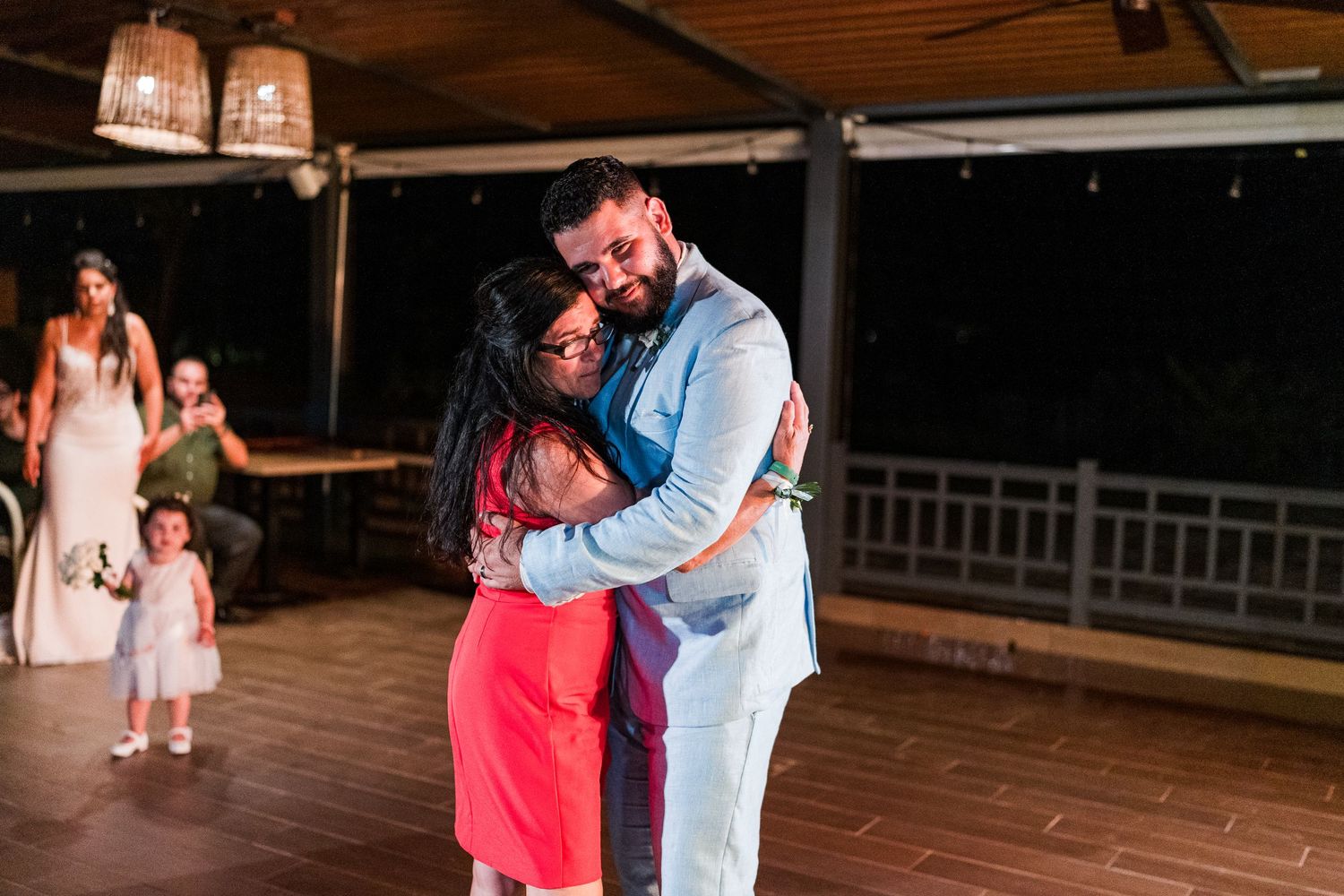 Groom tenderly embraces woman in red dress during emotional wedding reception dance with guests nearby.