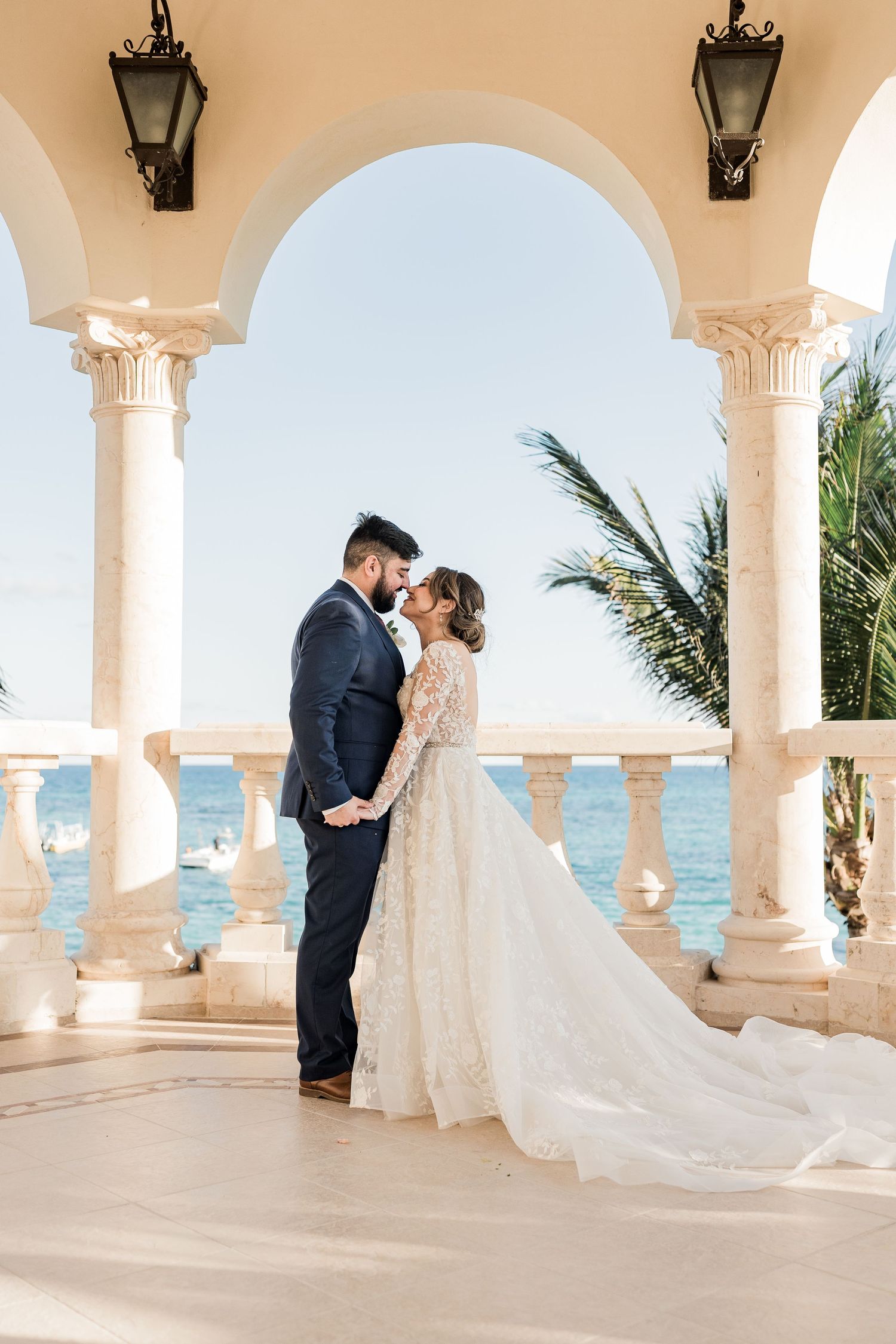 Bride and groom sharing romantic moment under arched colonnade with ocean and palm trees in background.