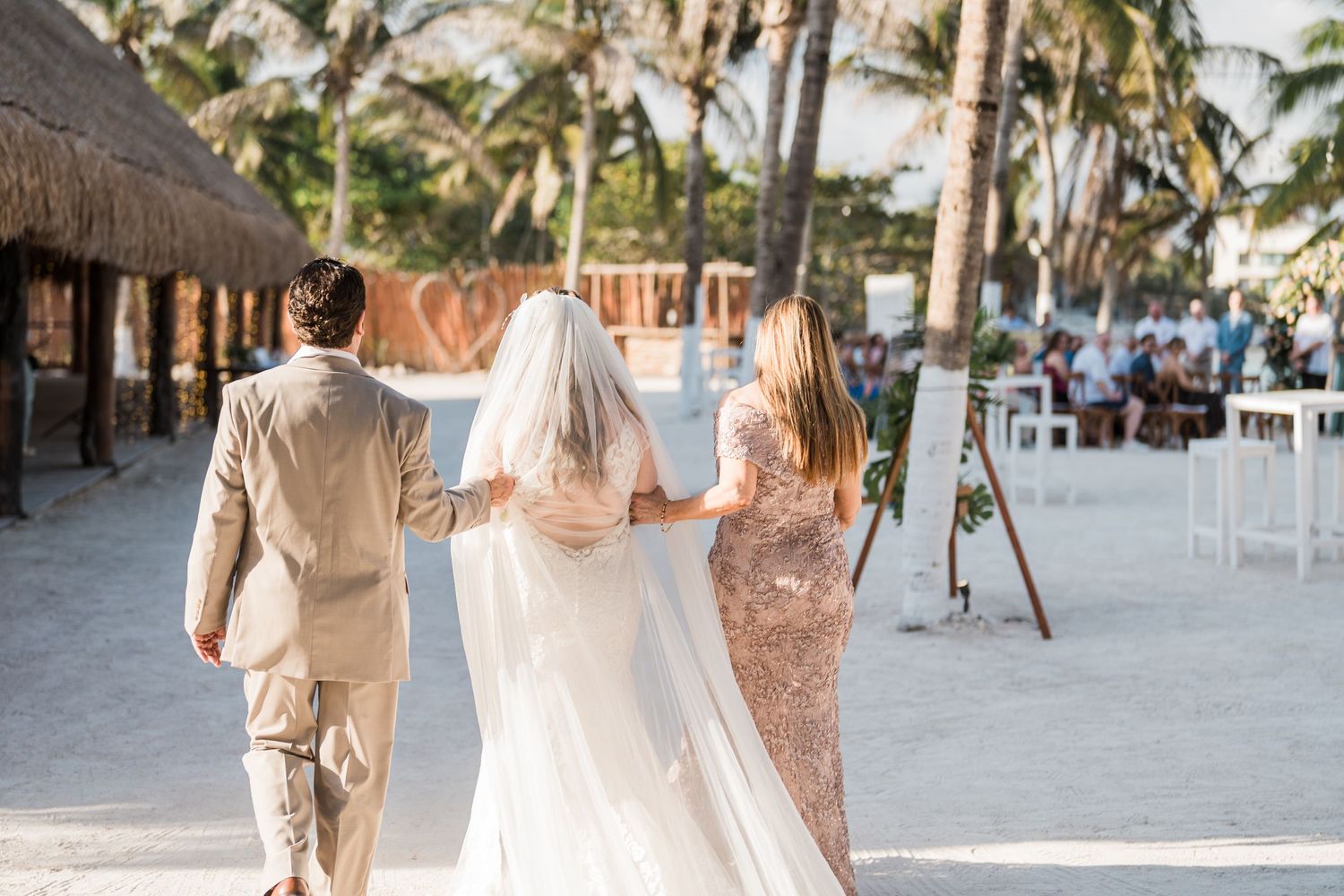 Bride walks down sandy beach aisle escorted by parents toward tropical wedding ceremony.