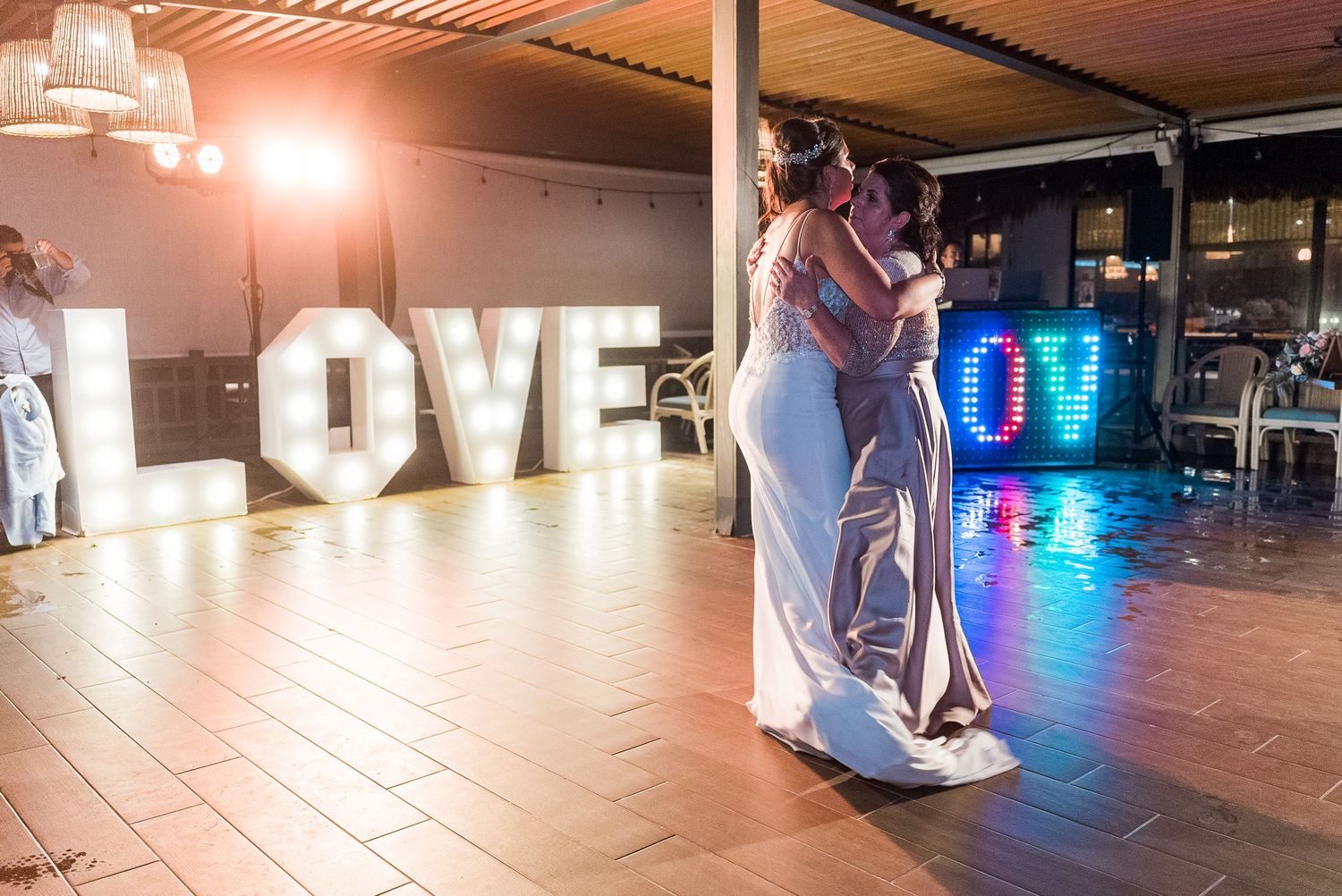 Bride shares an emotional first dance with a loved one near a glowing LOVE sign at reception.