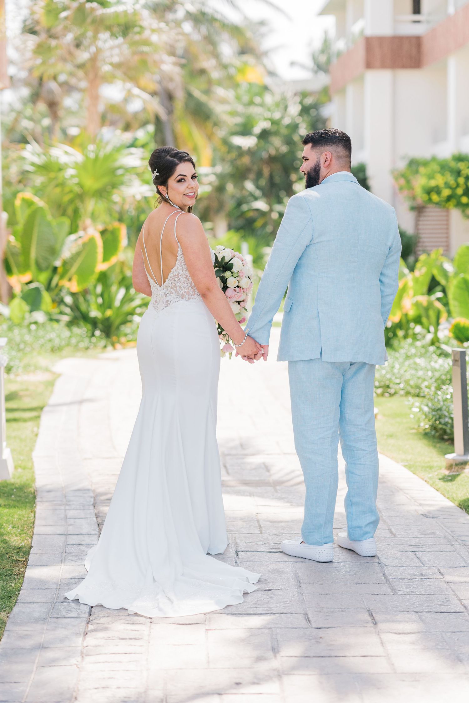 Bride and groom holding hands walking through tropical resort grounds, bride glancing back at camera.