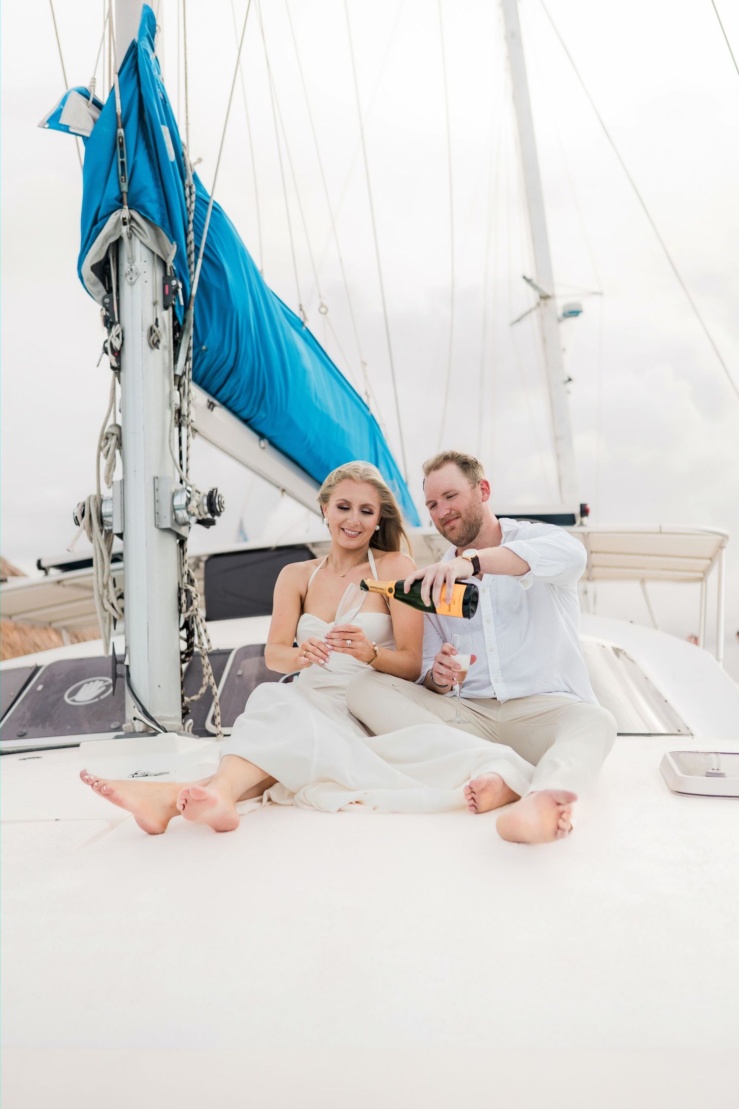 Cancun Engagement Photoshoot - Couple sitting on sailboat deck pouring champagne, wearing white outfits, barefoot, celebrating on the water.