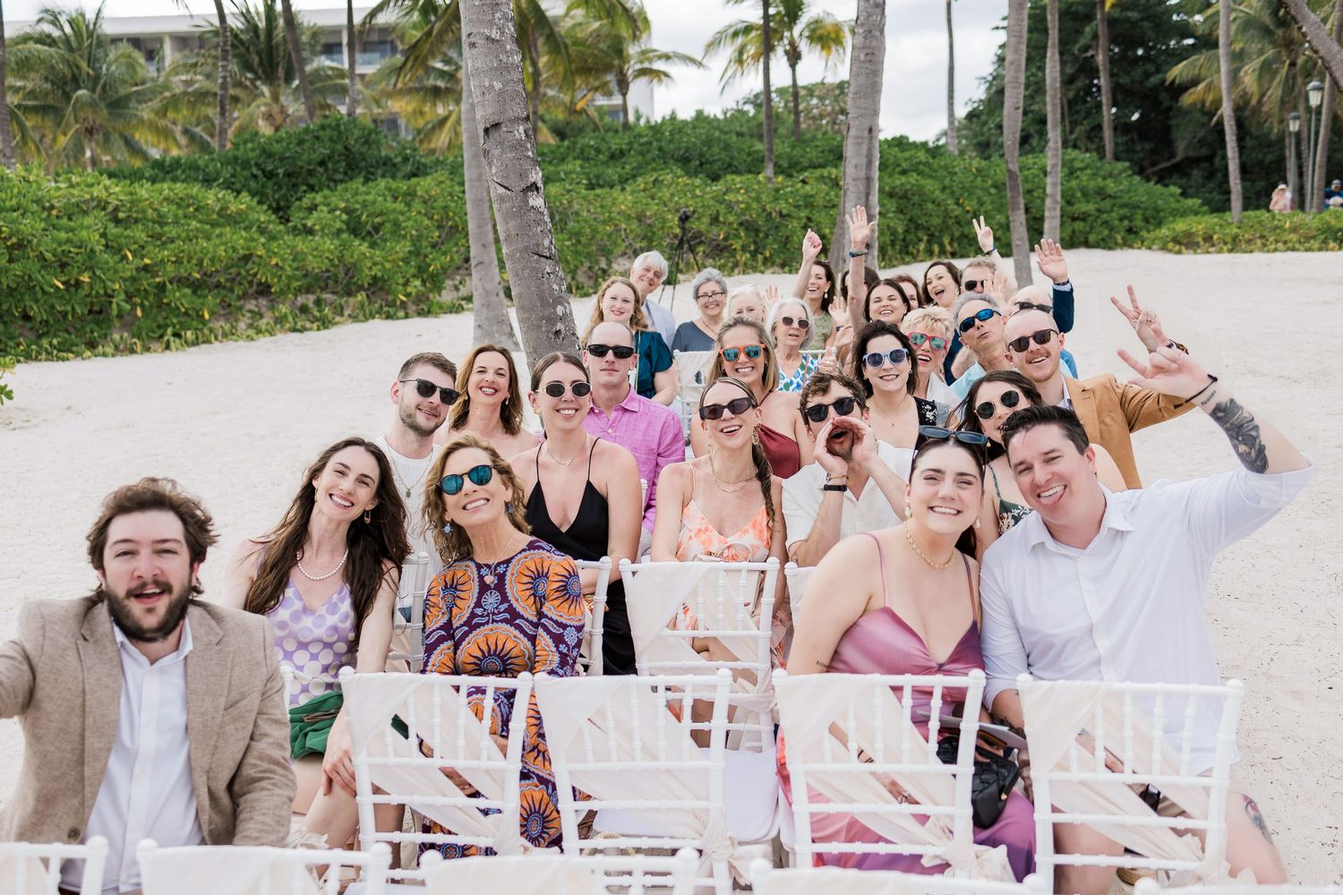 Large group of smiling wedding guests posing together on a tropical beach with palm trees and white chairs.