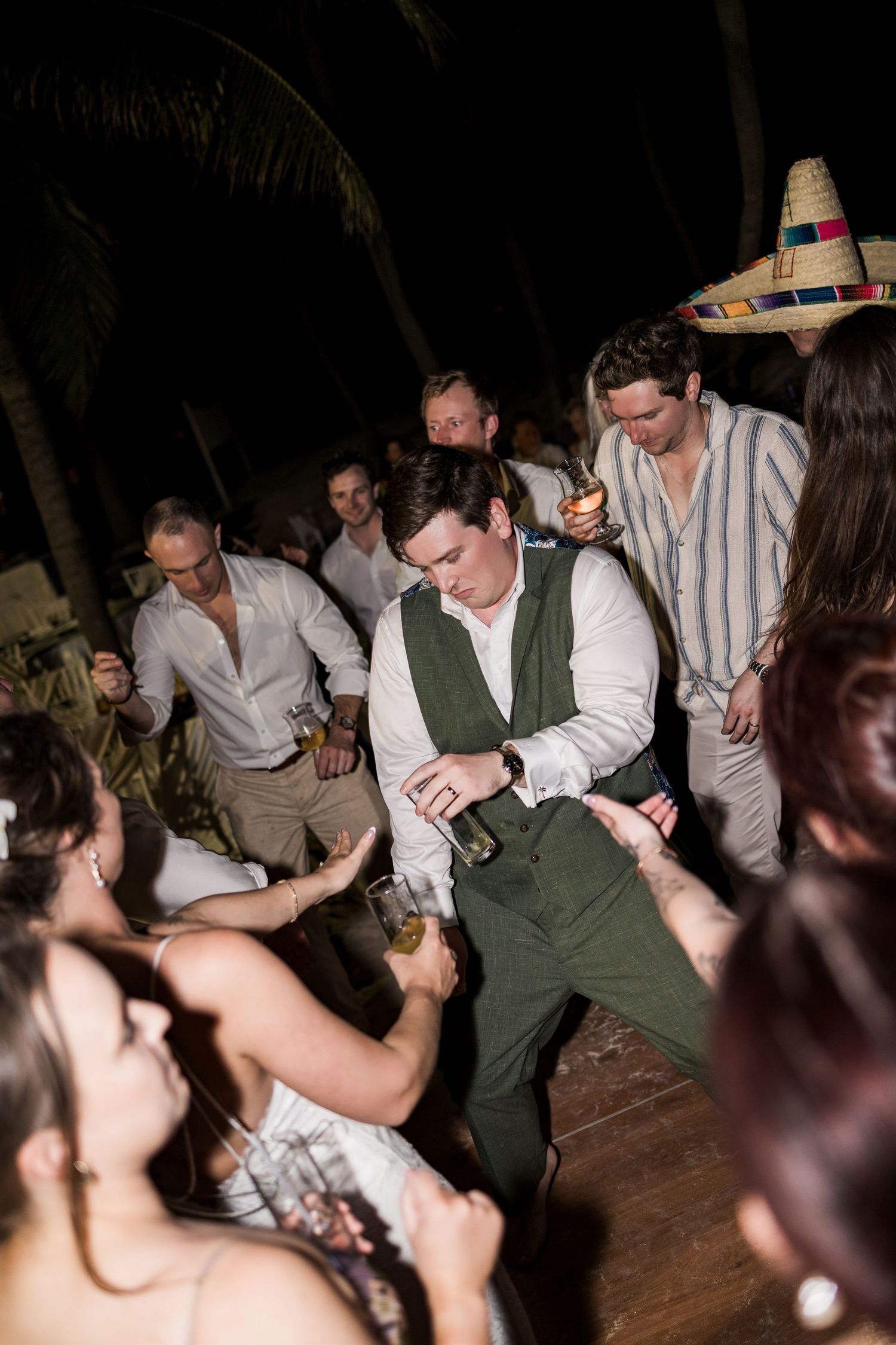 Guests dancing energetically at an outdoor wedding reception at night, with one man in a green vest holding a bottle.