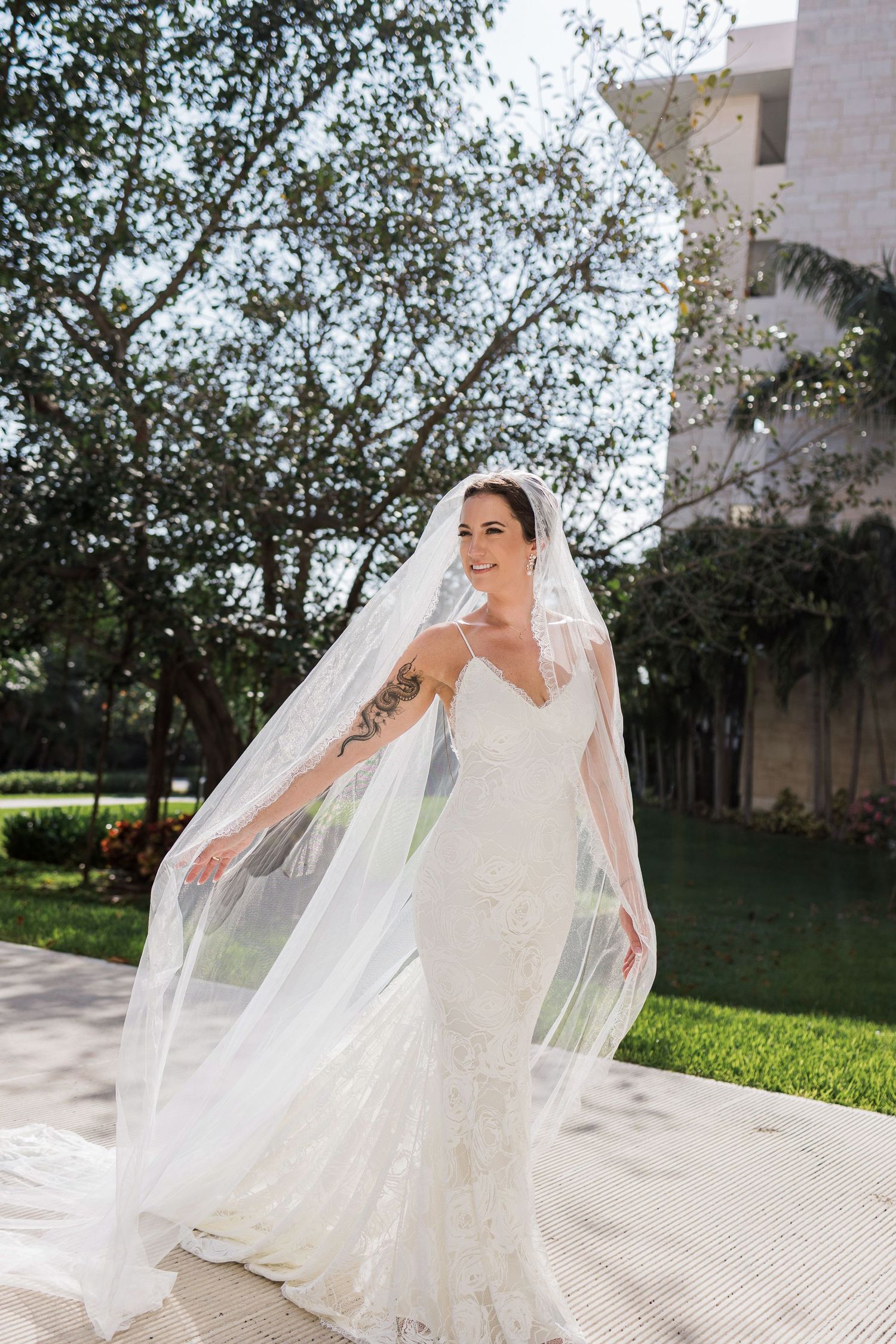 Bride in lace mermaid gown and flowing cathedral veil poses outdoors, smiling with arms outstretched on sunny day.