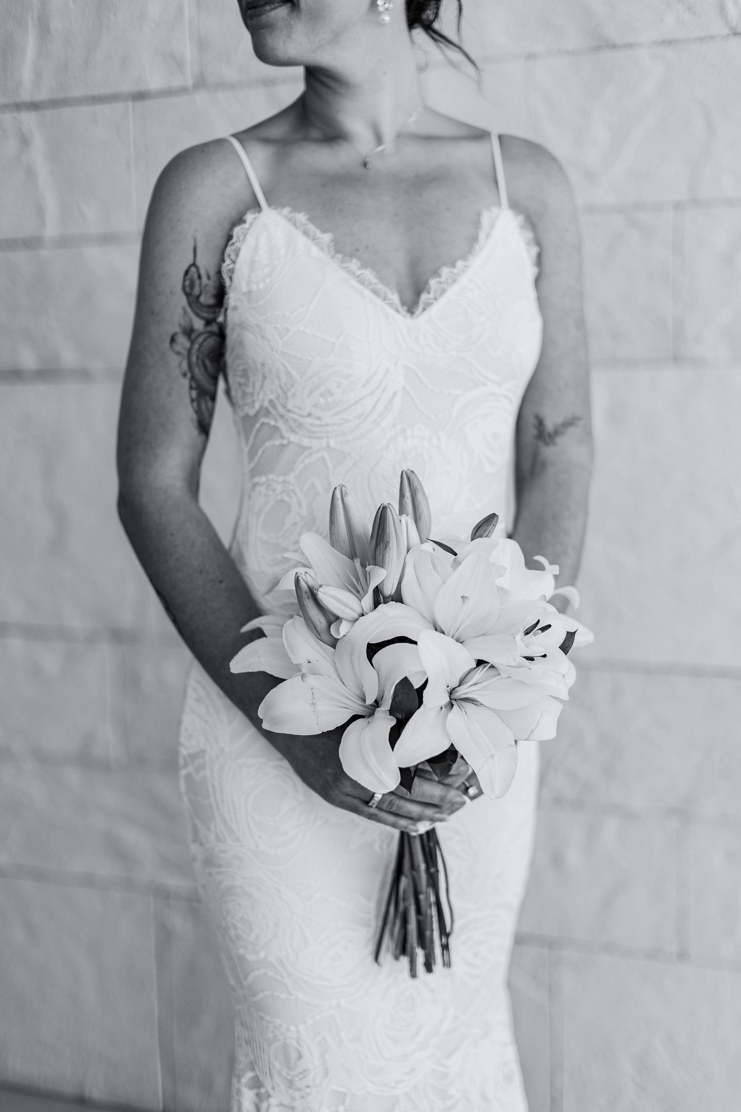 Bride in lace wedding gown holding lily bouquet, black and white photo showing tattoo on arm.