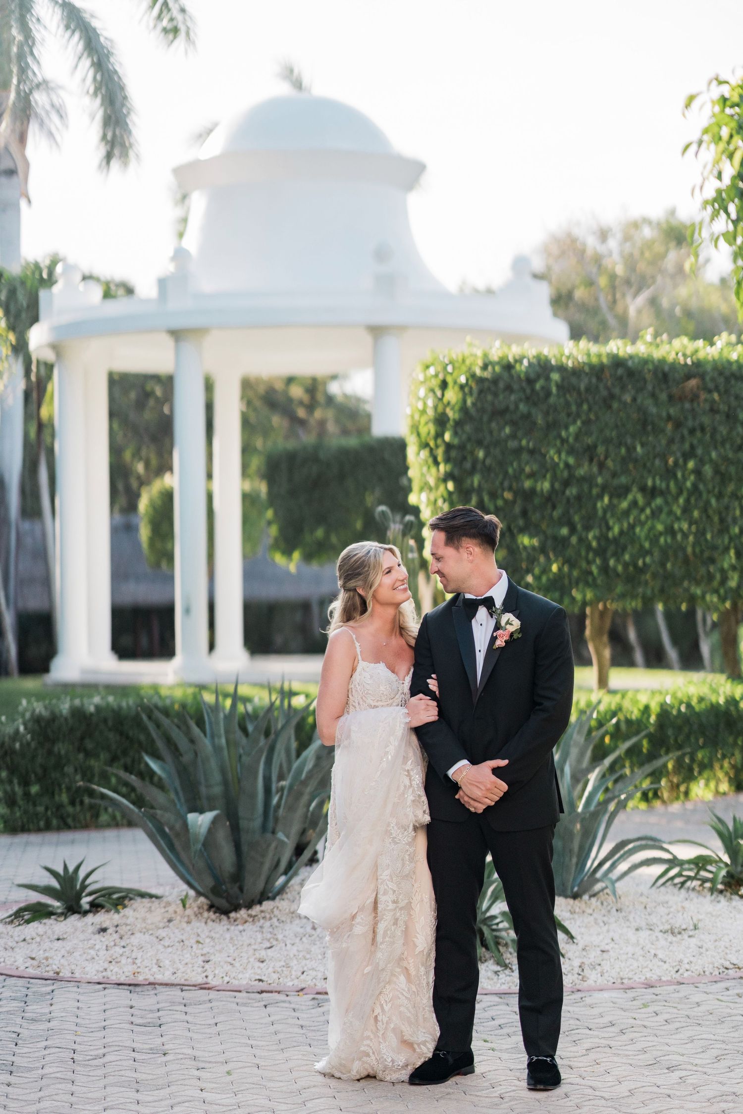 A bride and groom share a romantic moment in front of a white gazebo surrounded by tropical greenery on their wedding day.