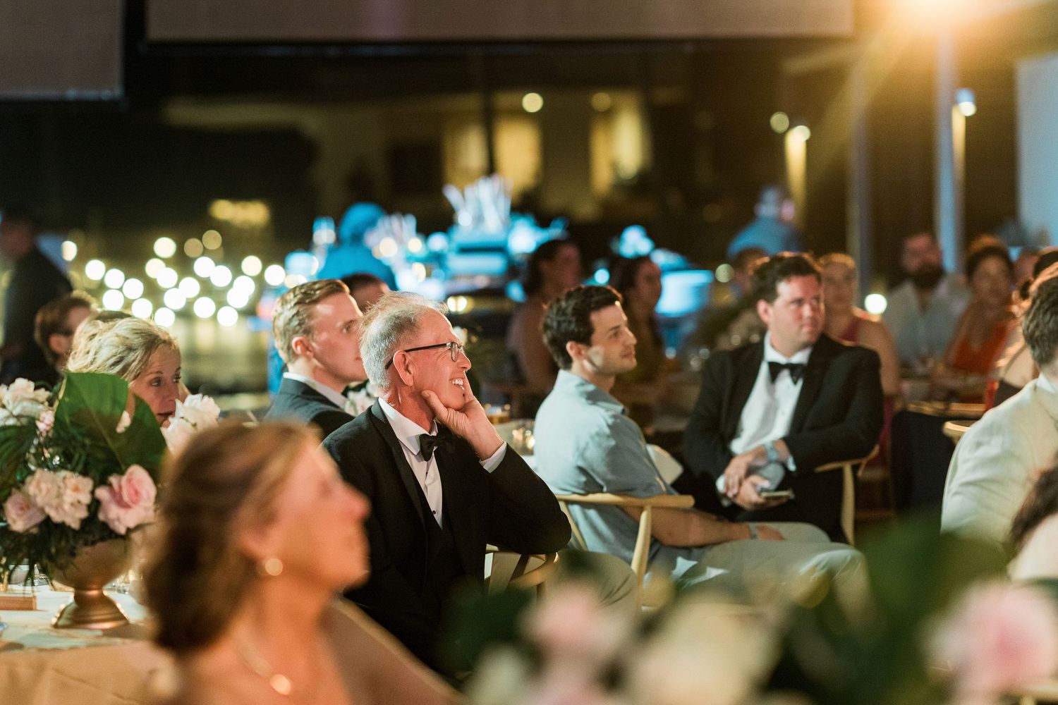 Elegantly dressed wedding guests seated at tables during a formal reception dinner in a warmly lit venue.