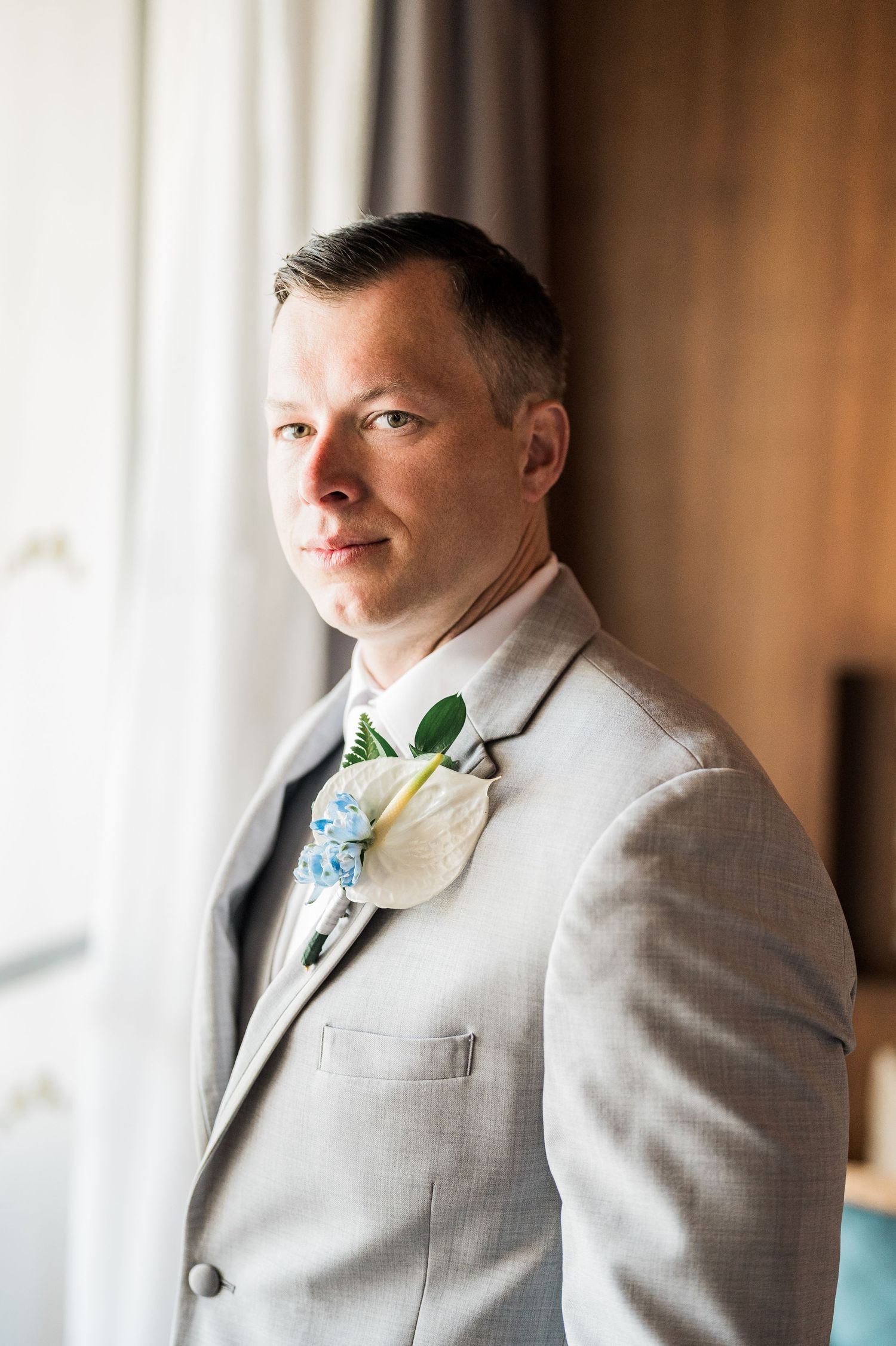 Groom in light gray suit with white and blue boutonniere poses near window on wedding day.
