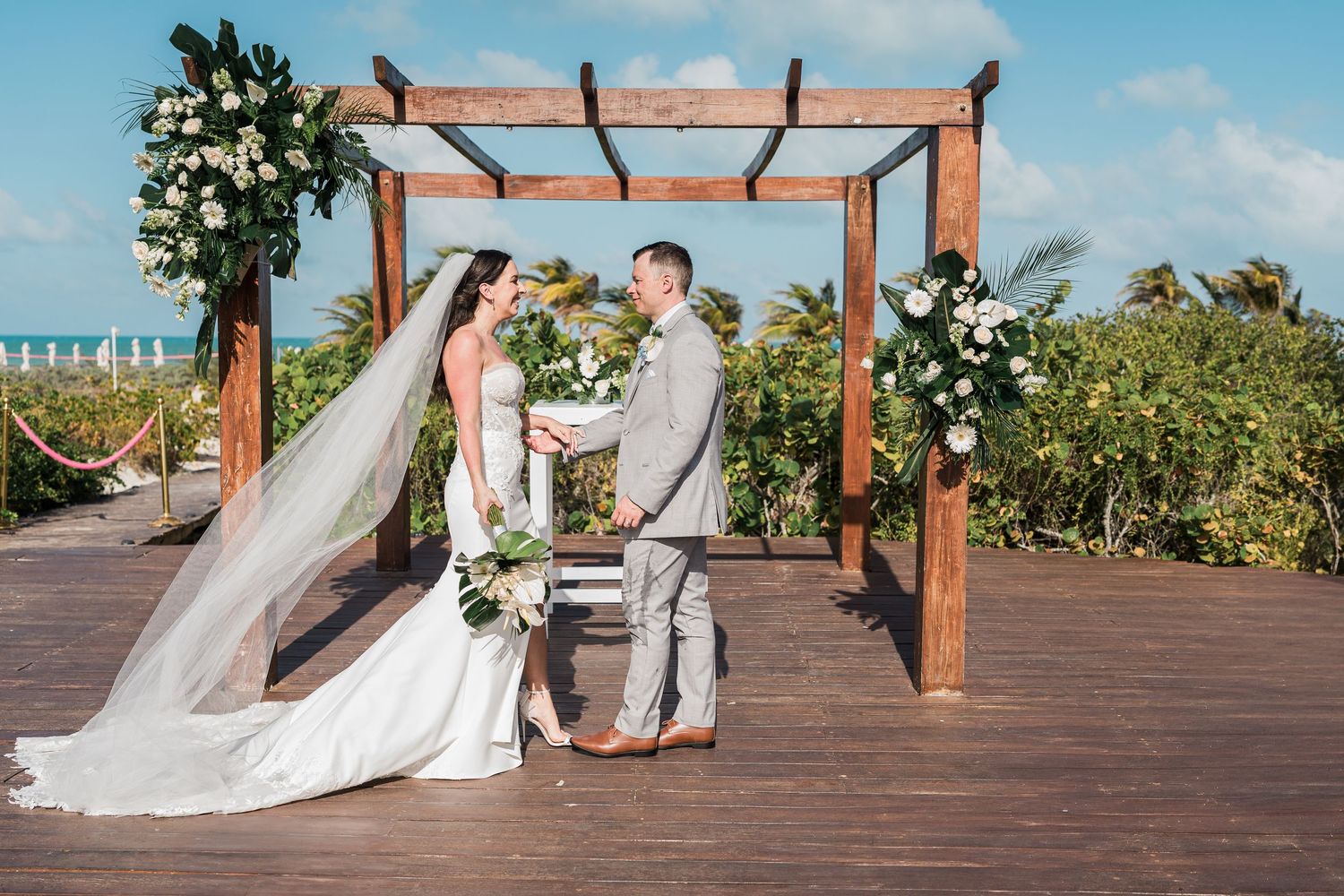 Bride and groom exchange vows under a wooden pergola adorned with white flowers on a tropical beachside deck.